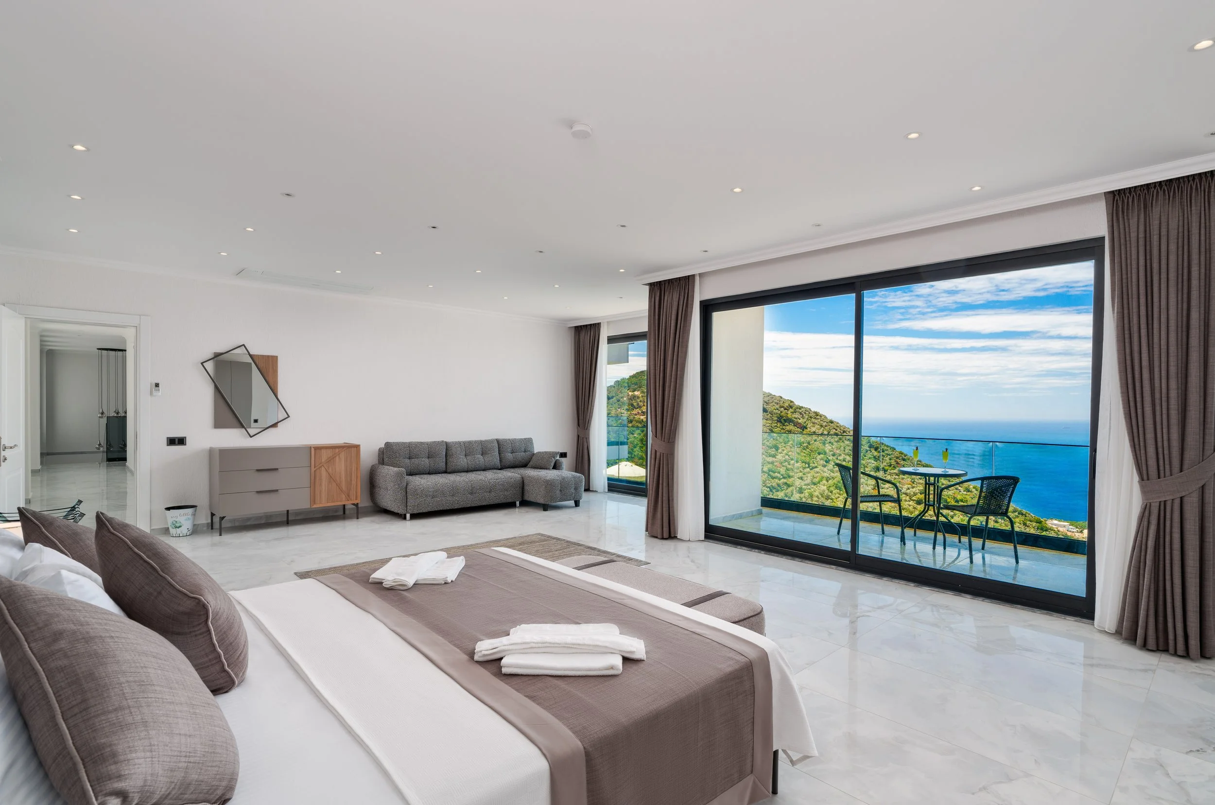 A modern hotel room with a large bed, sitting area, and a balcony view of greenery and the ocean.