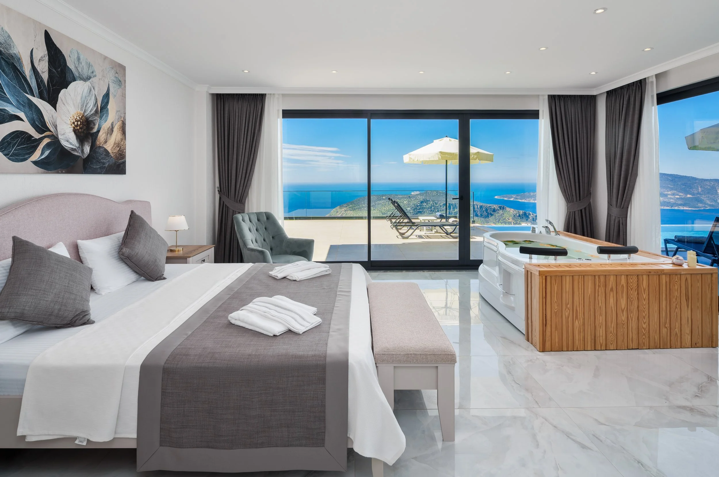 Hotel room with a large bed, pillows, bedside table with a lamp, and a view of the ocean and mountains through large sliding glass doors. There's a small deck with lounge chairs and an umbrella outside.
