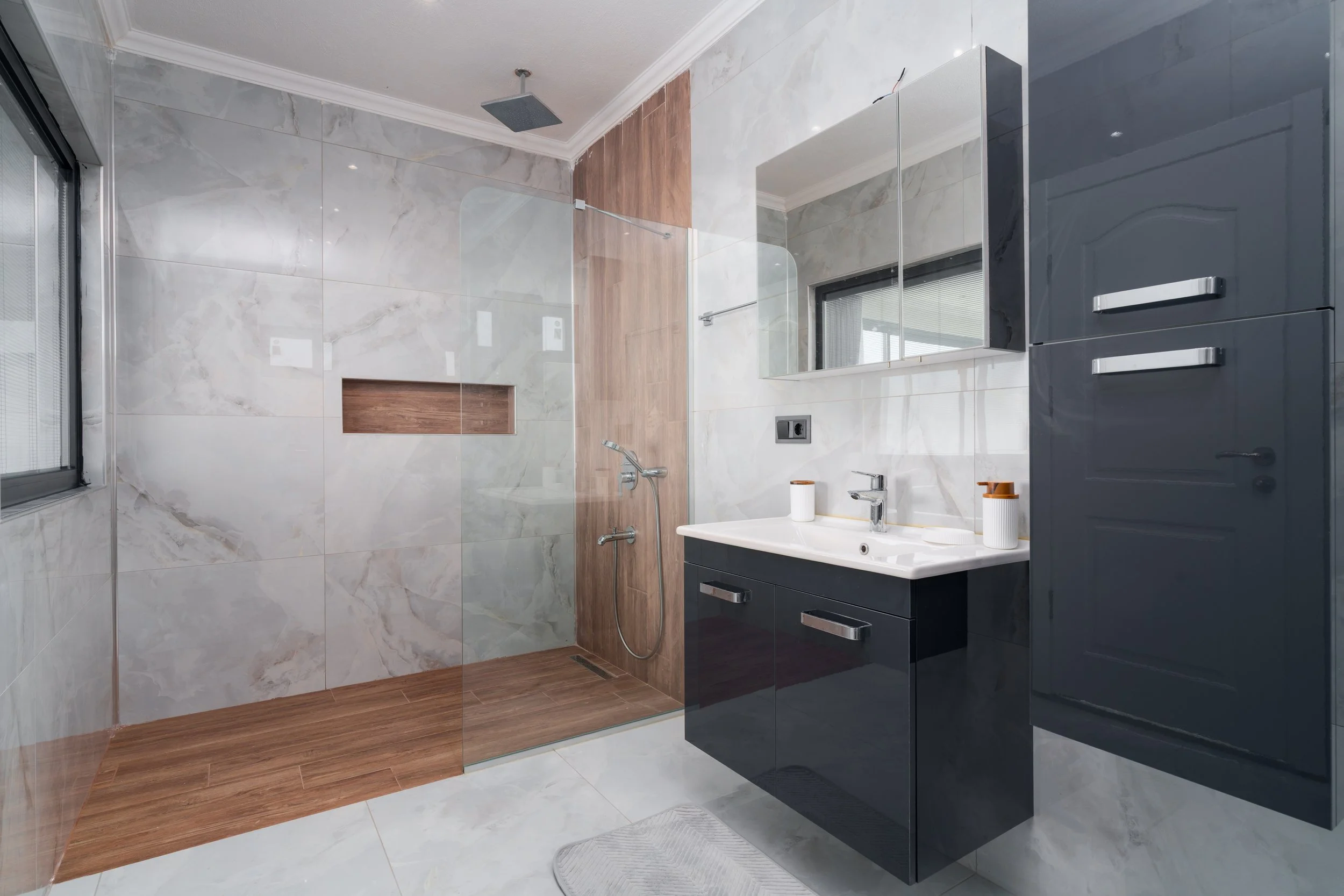 Modern bathroom with marble walls, wooden accents, a walk-in shower with glass partition, black cabinetry, and a small bathroom sink with a mirror and storage cabinets.