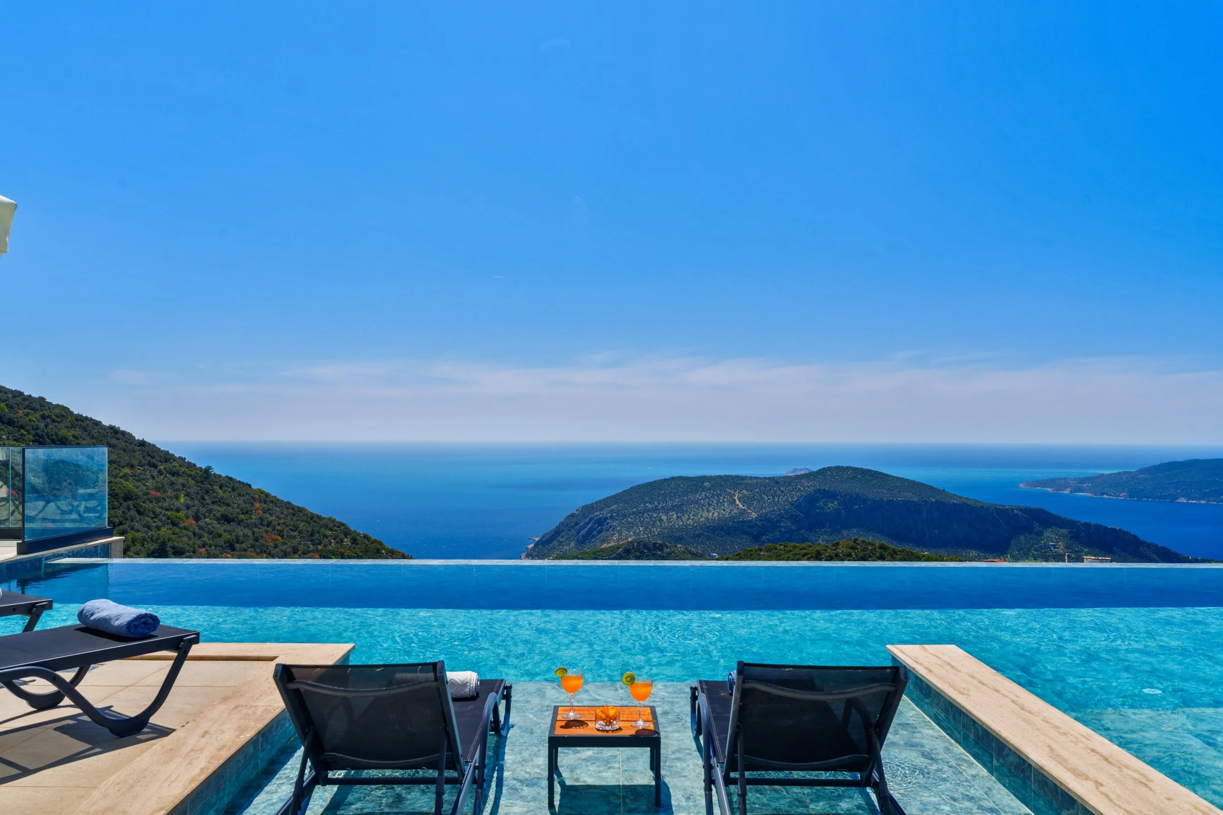 Swimming pool with lounge chairs and drinks overlooking a scenic view of hills and the ocean under a clear blue sky.