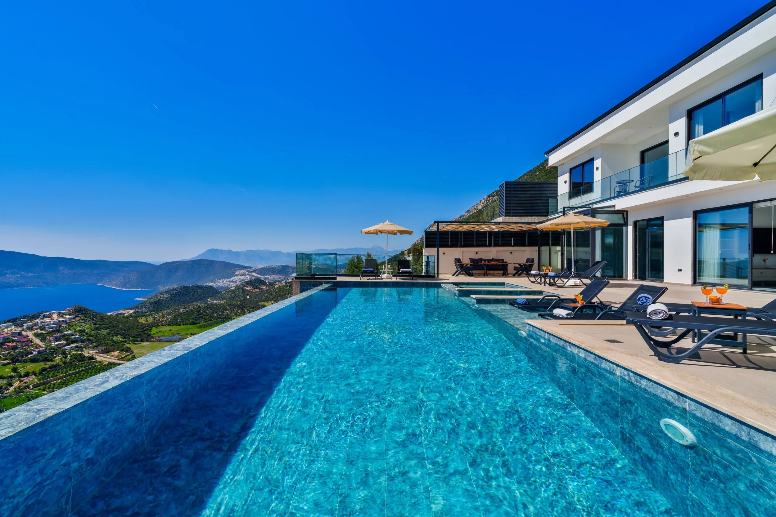 Modern white house with large glass windows and balcony, outdoor pool with lounge chairs and umbrellas, overlooking mountainous landscape and ocean under clear blue sky.