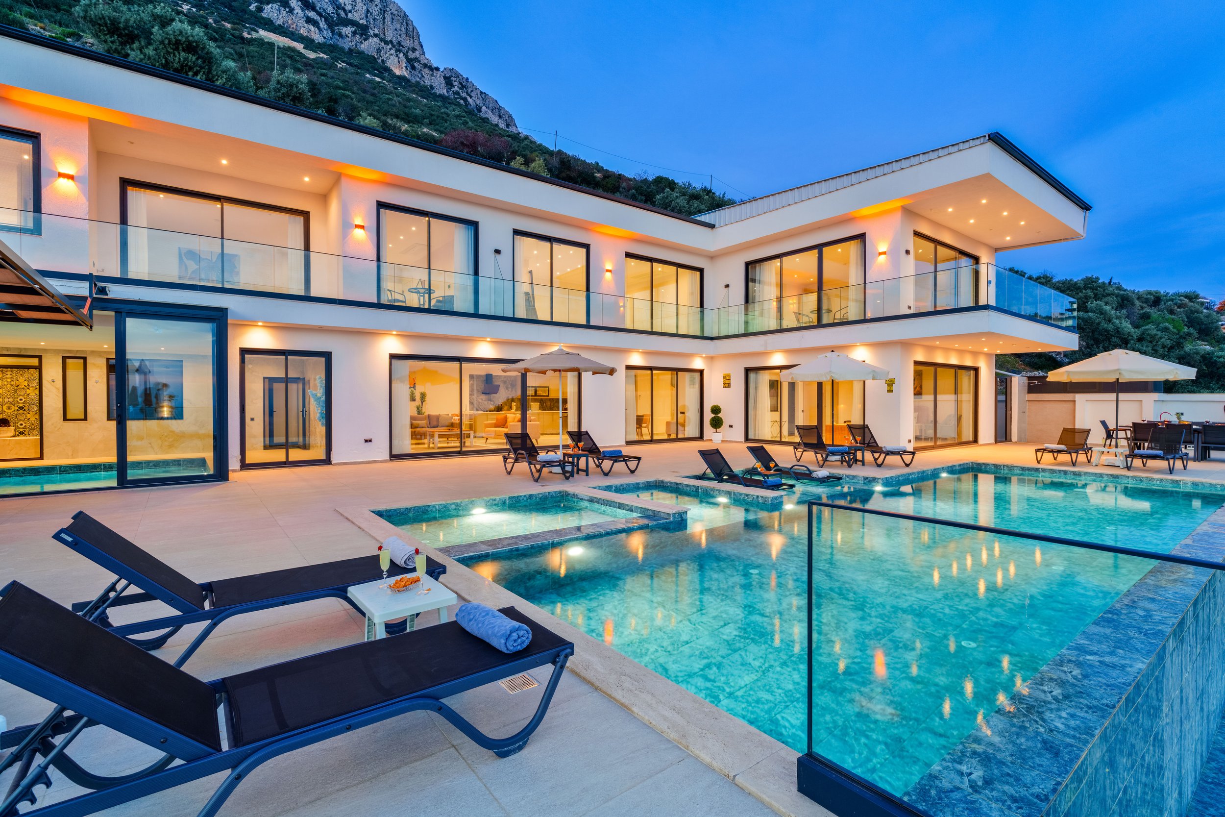 Modern two-story house with large glass windows and balcony, illuminated at dusk, with a swimming pool and lounge chairs in the backyard, surrounded by mountainous terrain.