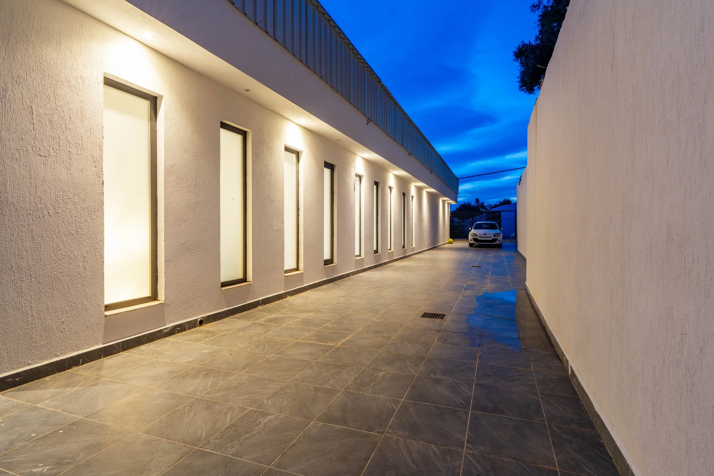 A modern building corridor in the evening with white exterior walls, illuminated by wall-mounted lights, and a parked white car at the end of the corridor under a dark blue sky.