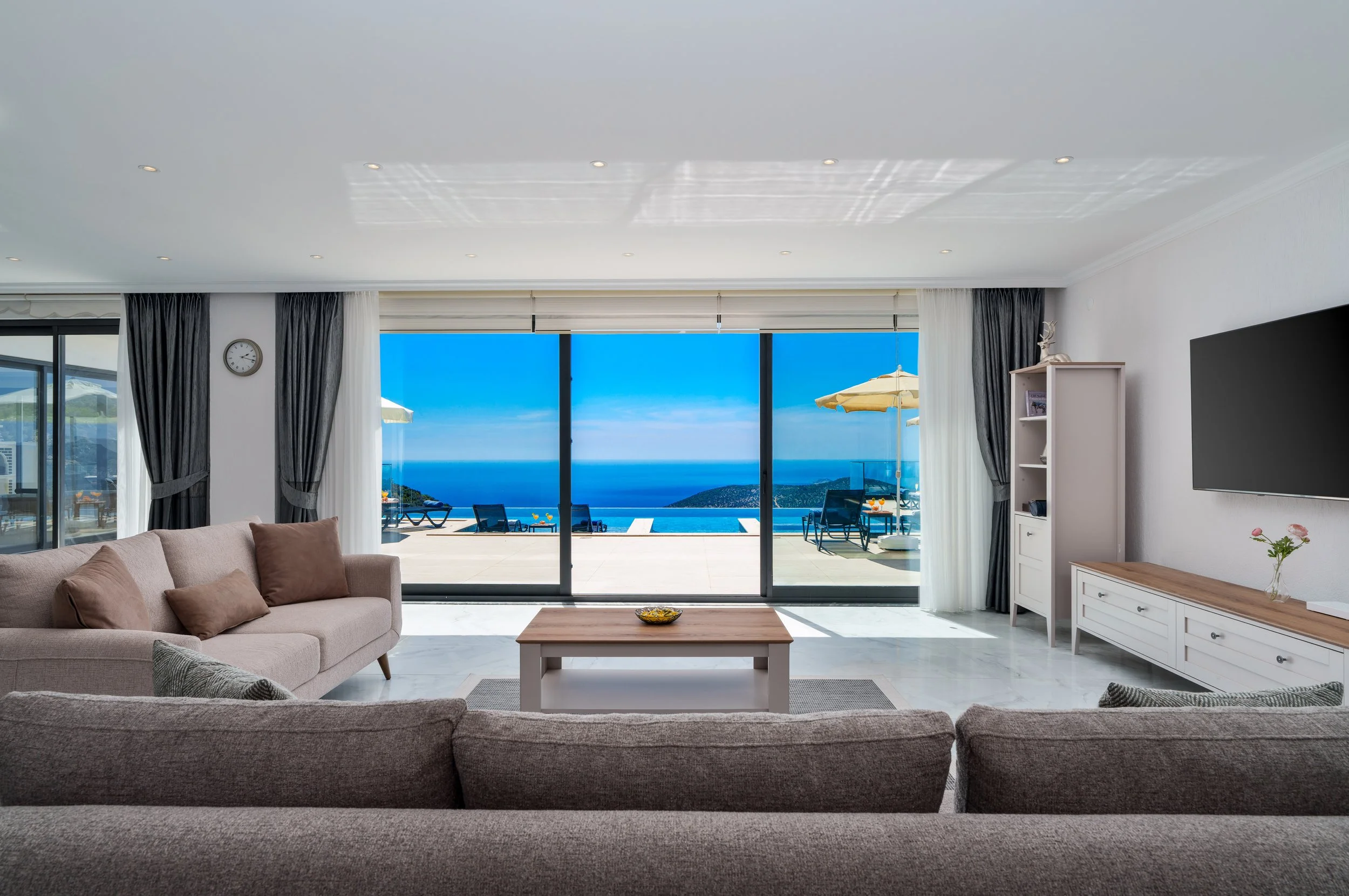 Living room with large glass doors leading to balcony with ocean view, beige sofa with brown pillows, wooden coffee table, white cabinet, TV, clock, and dark curtains.