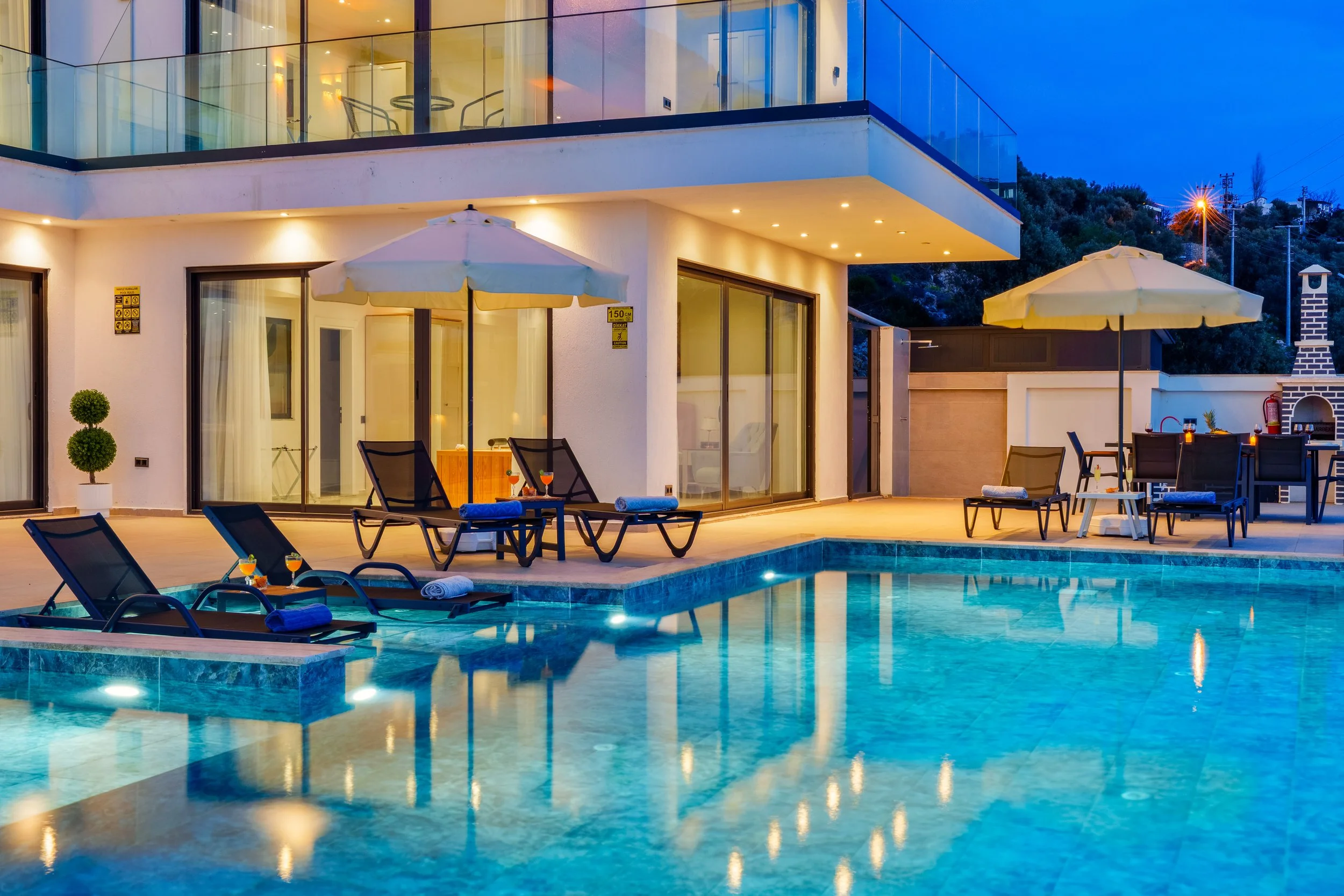 A modern house with a swimming pool and outdoor seating area, illuminated at night. The house has large glass windows and balconies, with umbrellas and lounge chairs around the pool.