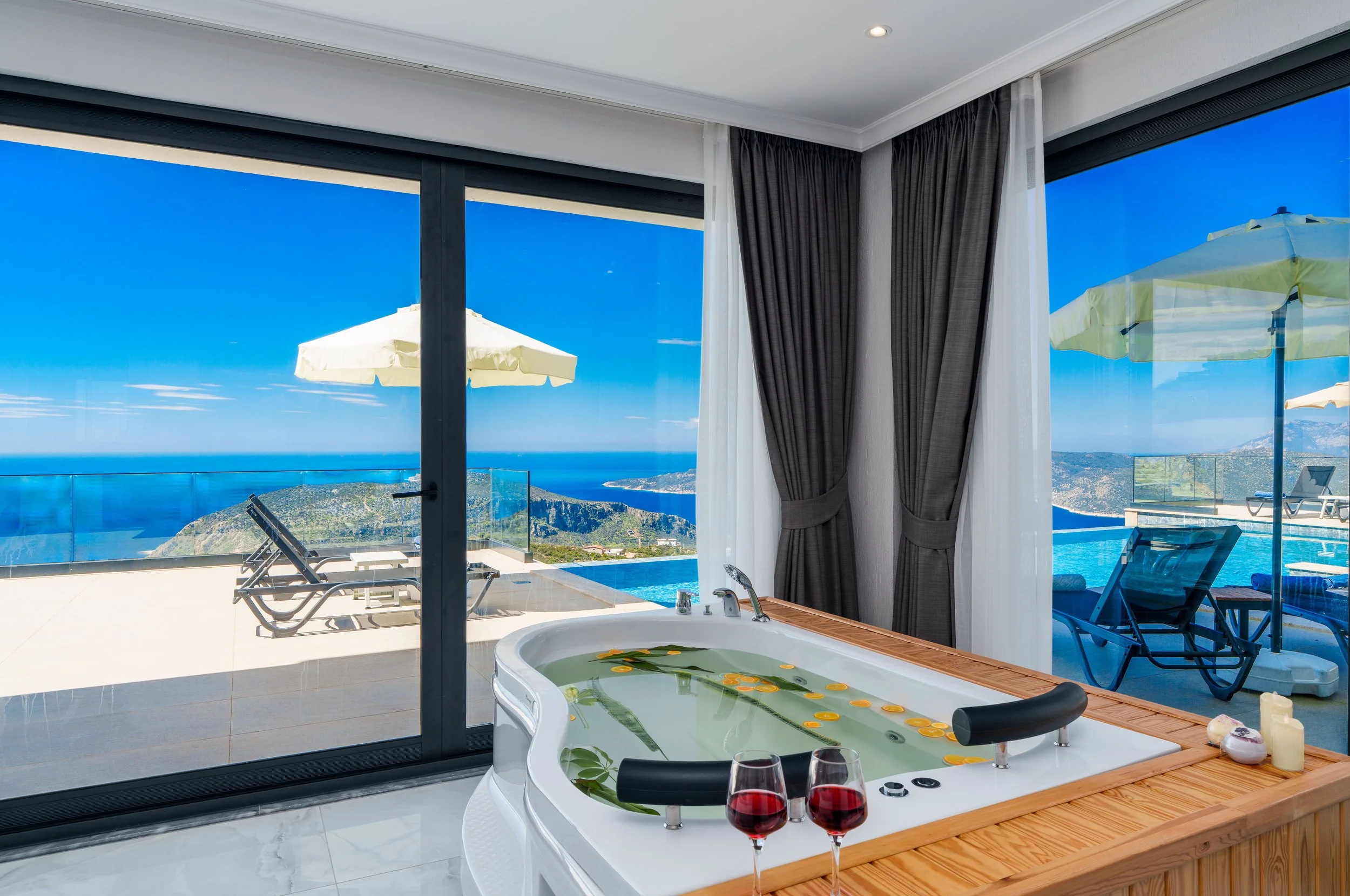 A luxurious bathroom with a view of a swimming pool, patio furniture, parasols, and scenic landscape with mountains and the ocean through large glass sliding doors. Inside, there is a whirlpool bathtub with rose petals and two glasses of red wine on 