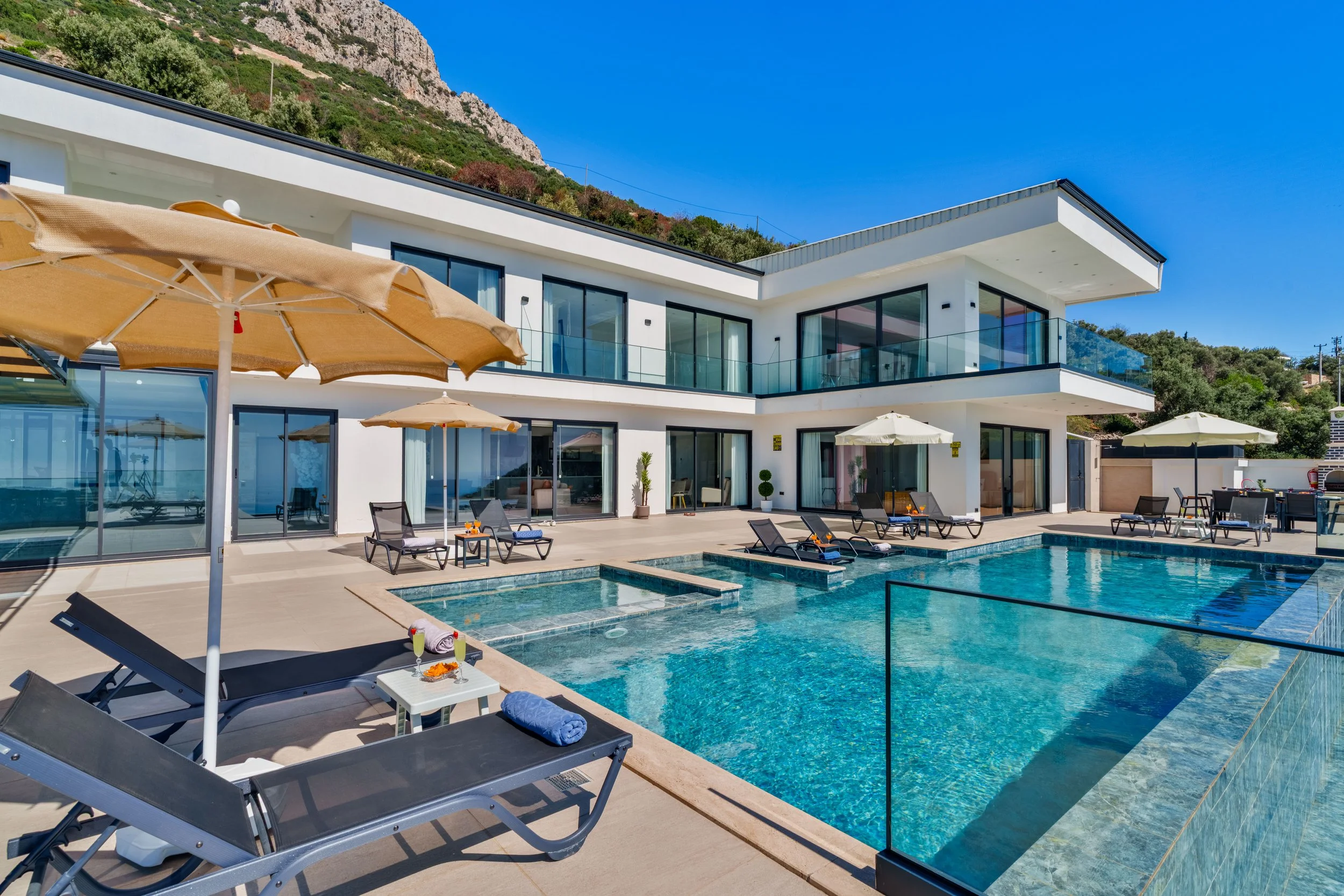 A modern two-story house with large glass windows, balconies, and a patio area with lounge chairs and umbrellas. There is a swimming pool in the foreground and a hillside with greenery in the background under a clear blue sky.