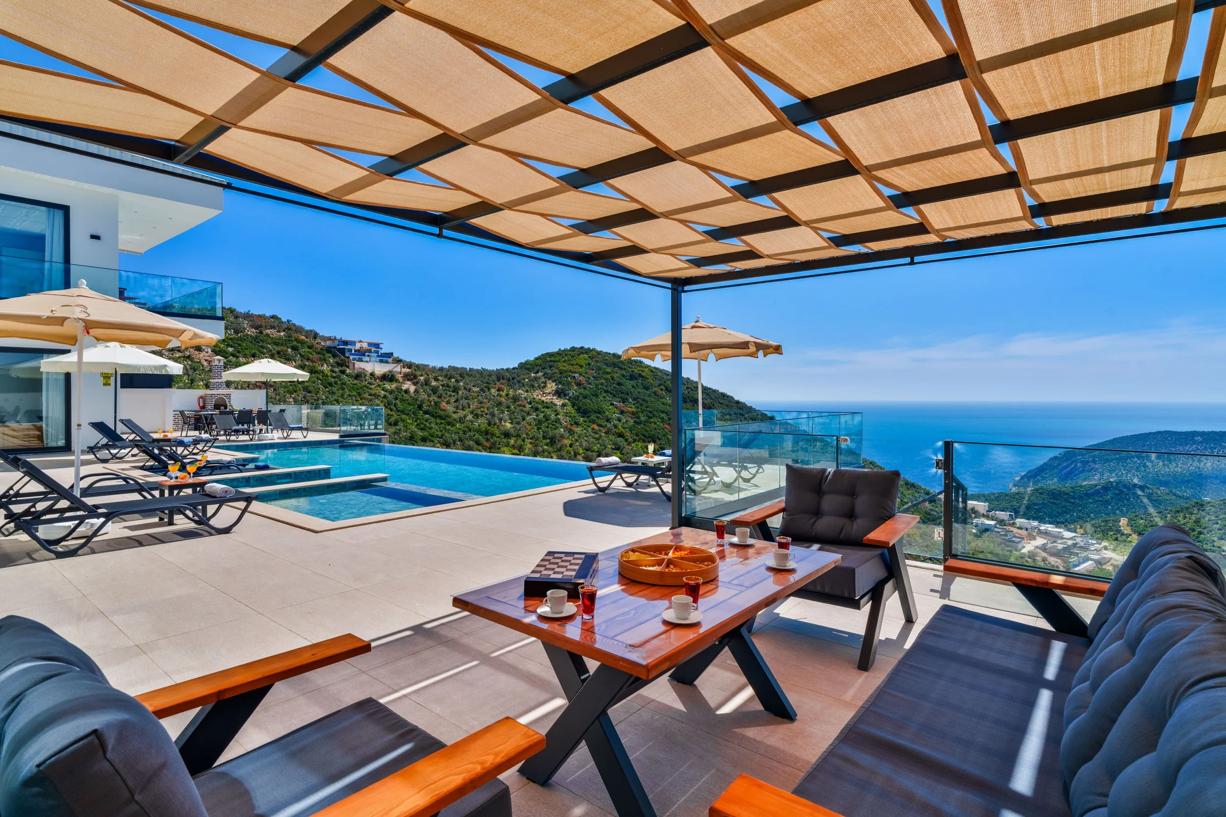 View of an outdoor terrace with lounge chairs, tables with breakfast settings, umbrellas, a swimming pool, a modern building, and hilly landscape with clear blue sky.