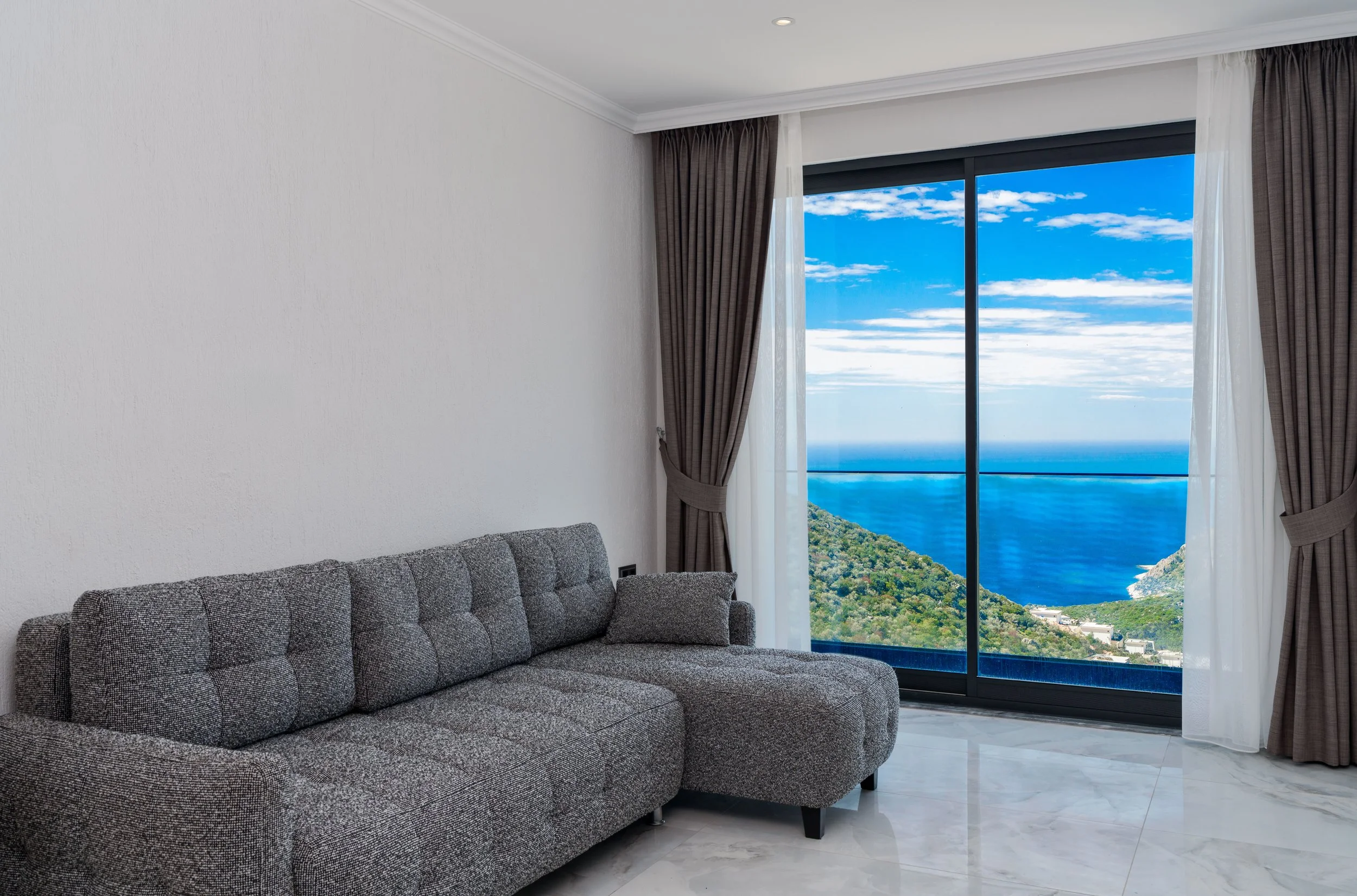 A living room with a gray upholstered couch, white walls, marble floor, and a large window with black and white curtains showing a view of blue ocean, green hills, and a partly cloudy sky.