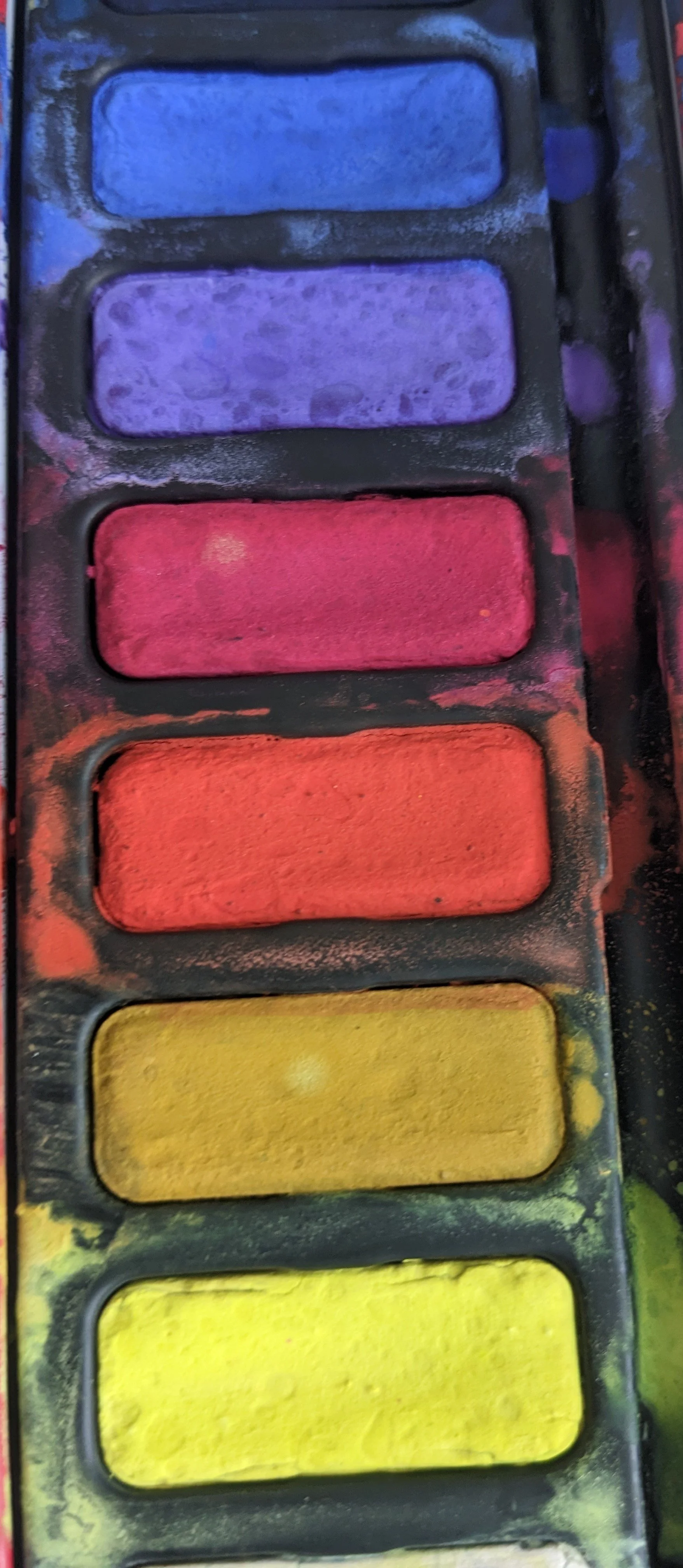 A watercolor paint palette with six rectangular pans of colors: blue, purple, red, orange, yellow, and green, with some colors mixed and some chalky residue around the pans.