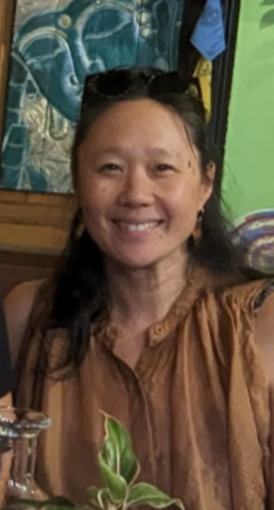 Smiling woman with long dark hair, sunglasses on head, wearing a brown top, in a colorful setting with artwork and green wall in the background.