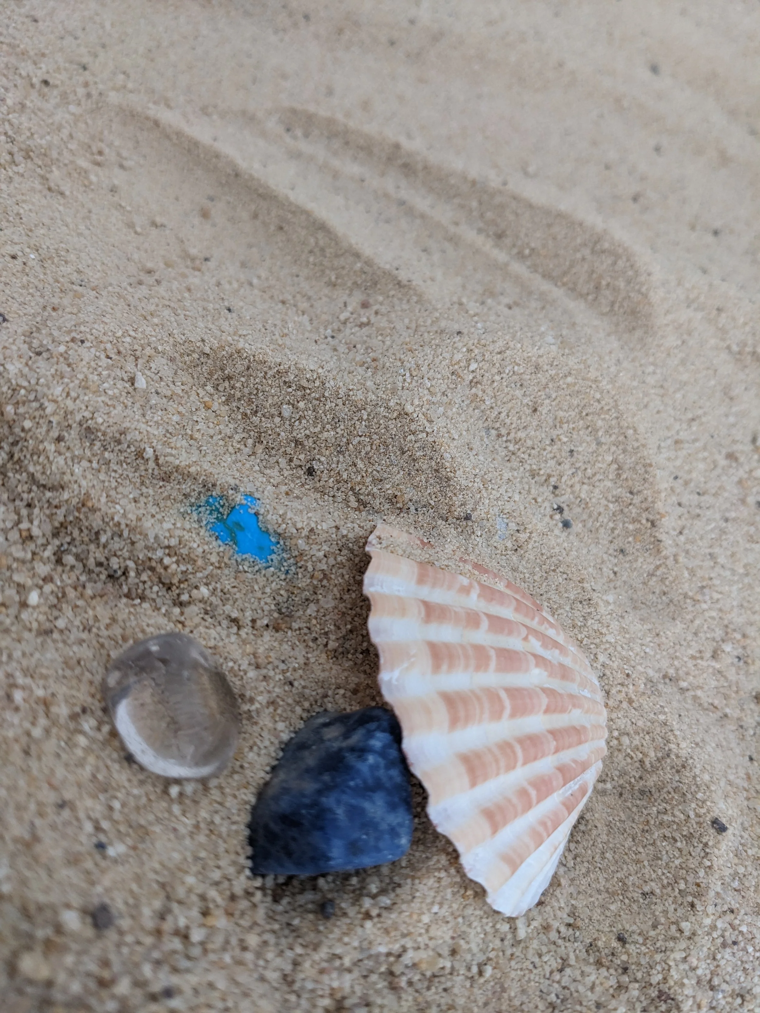 A sandy beach with a striped seashell, a small blue item, a clear glass orb, and black rocks. A person's foot in a sandal is at the bottom of the image.