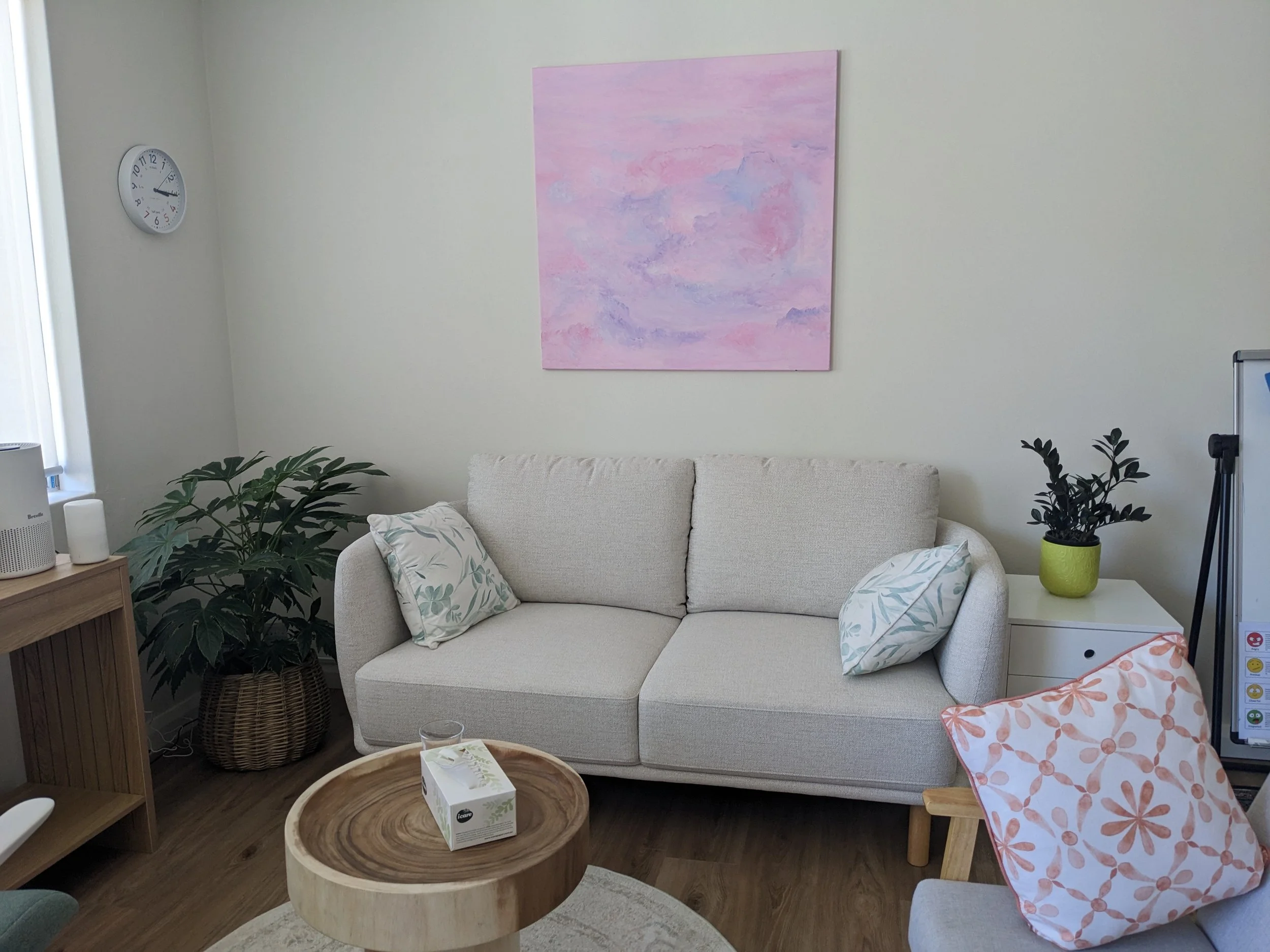Living room with a white couch, pink and white pillows, a wooden coffee table with tissue box, houseplants, a wall clock, and a pink abstract painting.