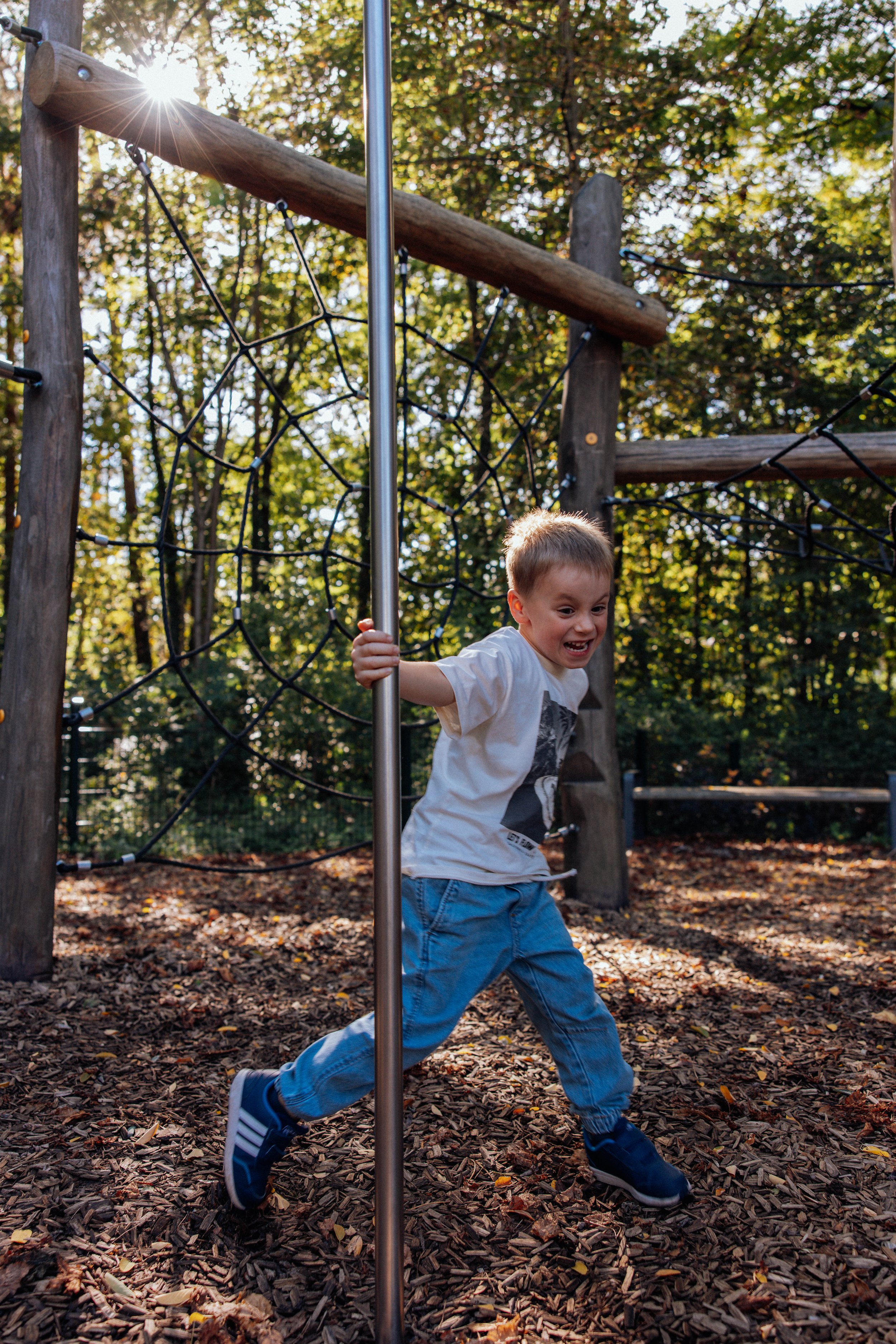 Junge läuft lachend um einen Spielpfosten in einem Spielplatz im Freien, umgeben von Bäumen mit Herbstlaub auf dem Boden.