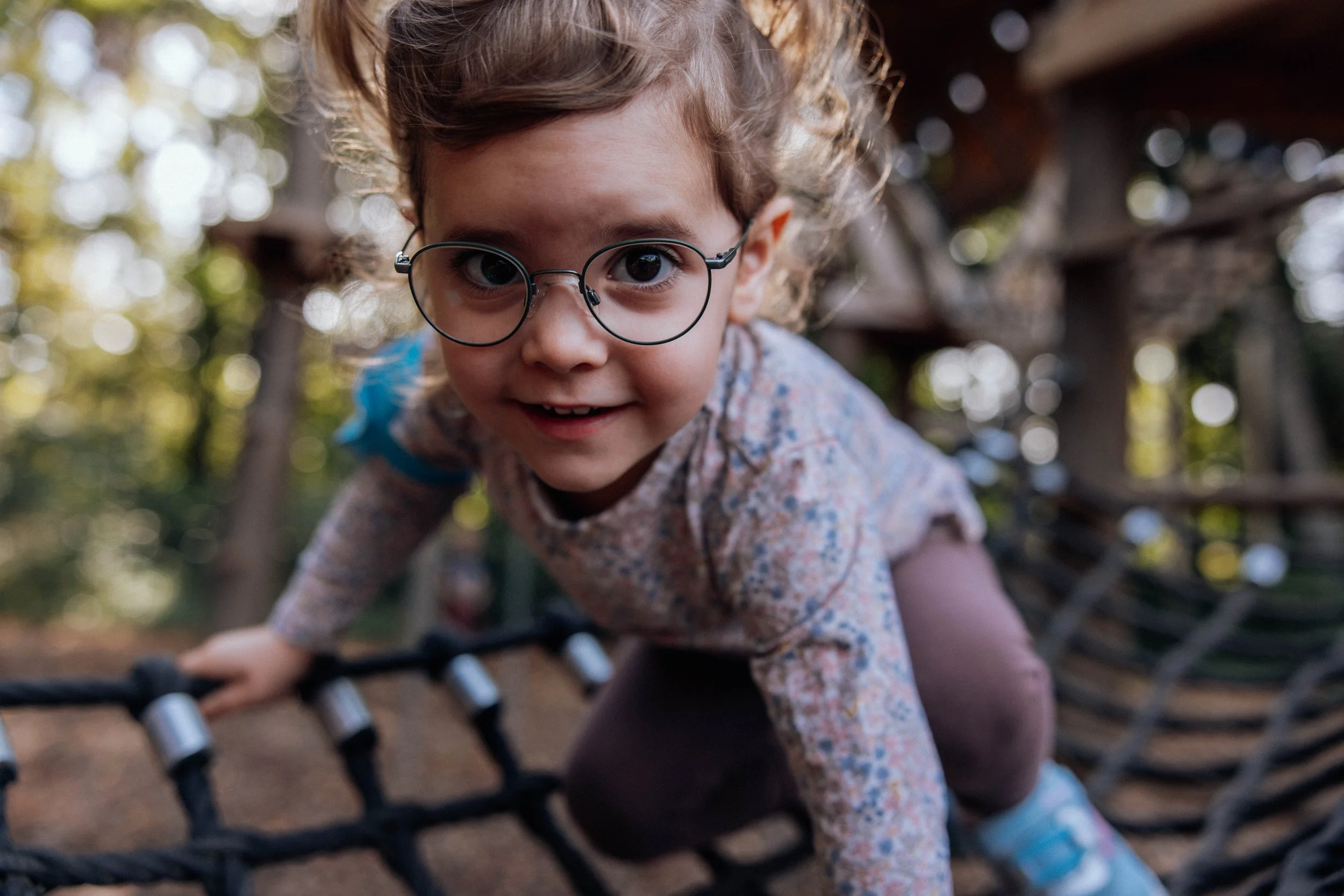 Ein junges Mädchen mit runden Brillen und braunen, lockigen Haaren klettert auf einem Holzspielplatz im Freien, umgeben von grünen Blättern und Sonnenlicht.