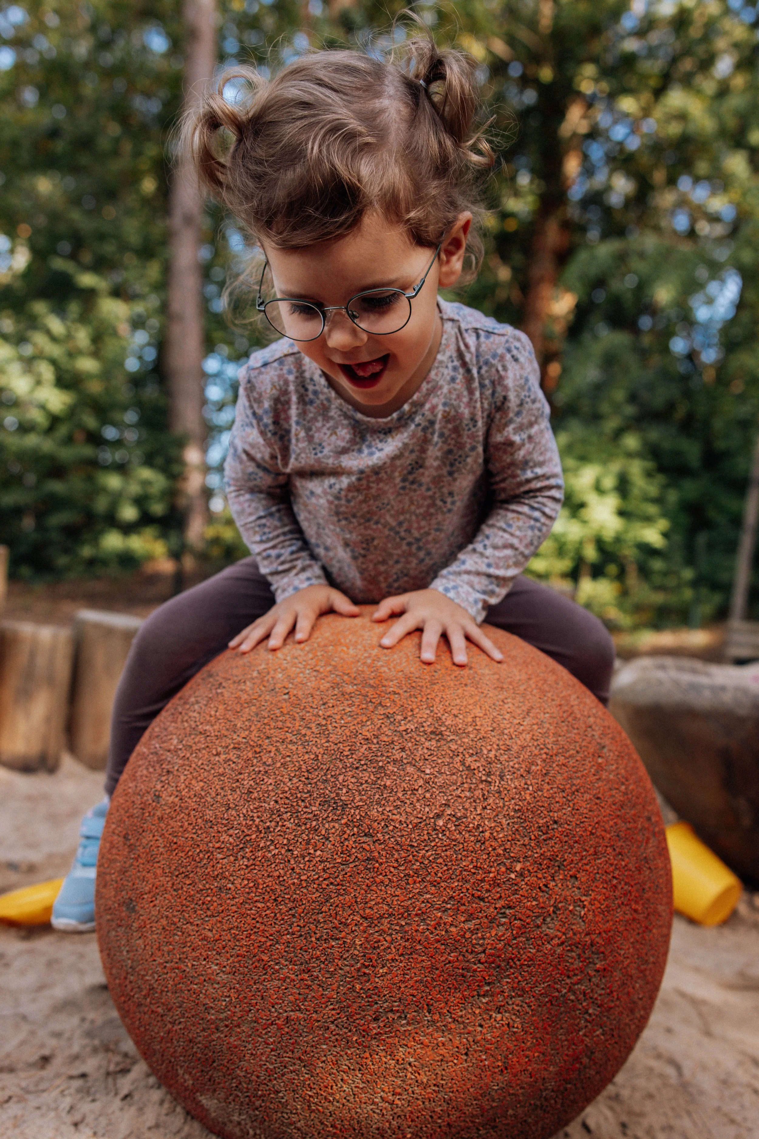 Ein kleines Mädchen mit Brille und blondem, lockigem Haar spielt draußen auf einem Spielplatz. Sie sitzt auf einem großen, orangefarbenen, kugelförmigen Spielgerät aus Gummi.