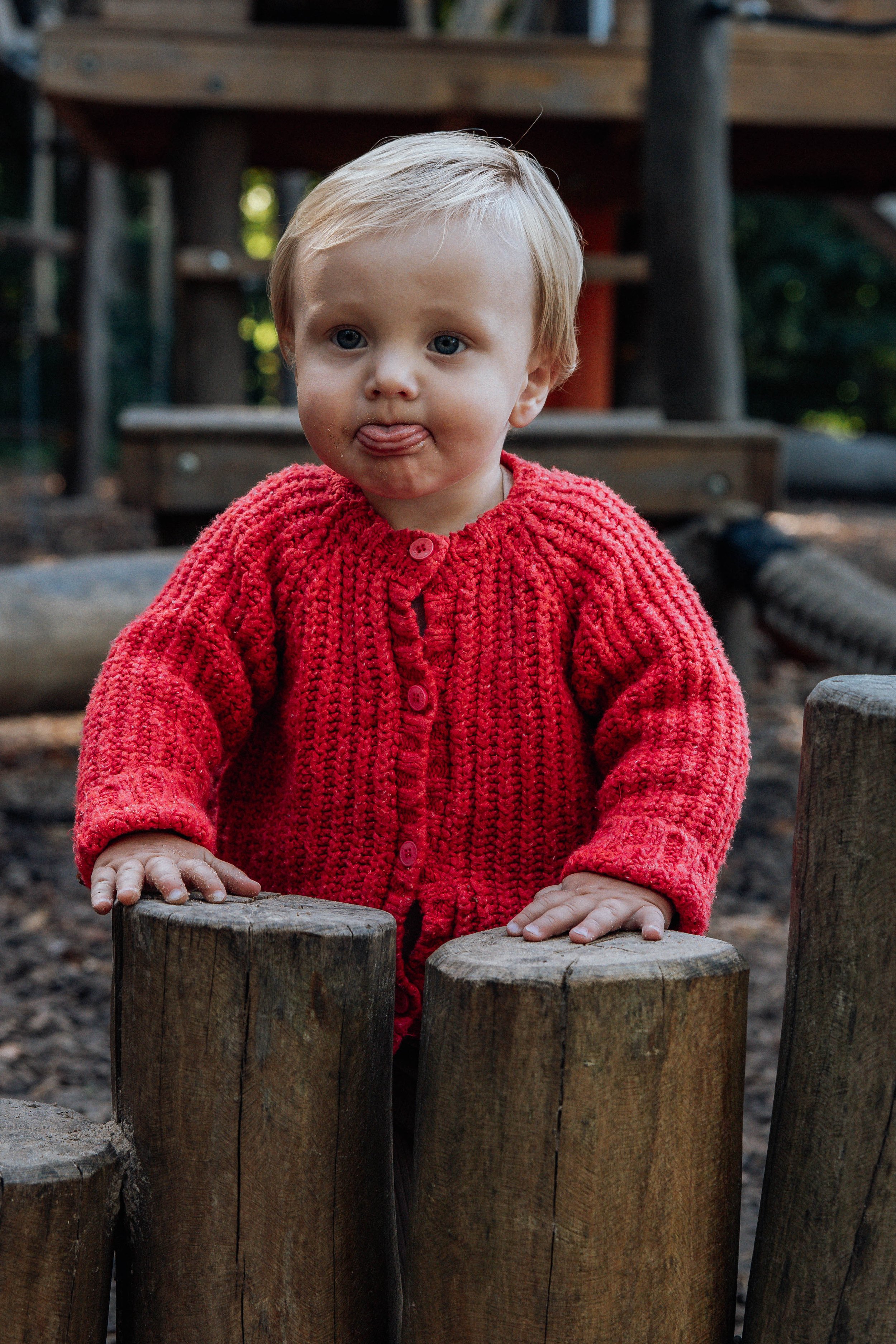 Ein kleiner Junge mit blonden Haaren trägt eine rote gestrickte Jacke und steckt die Zunge heraus. Er steht hinter einem Holzzaun und hält sich an den Baumstämmen fest.