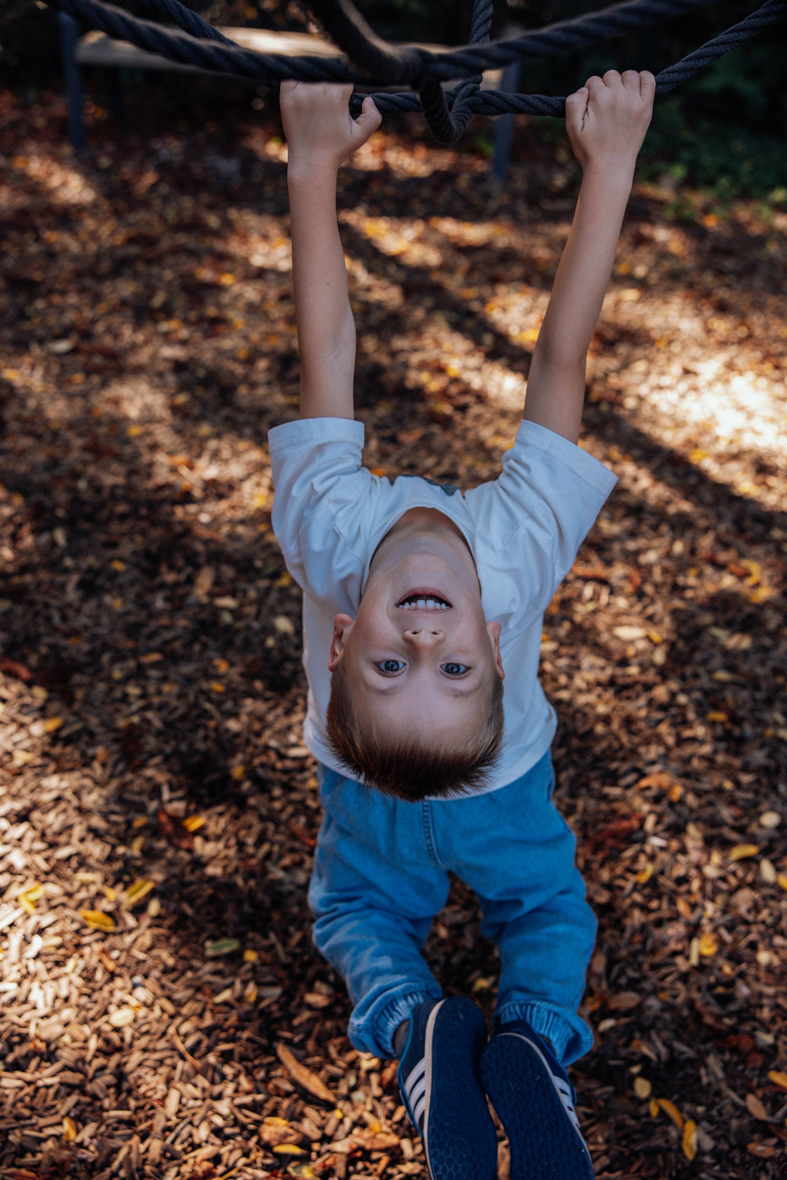 Ein Kind hängt an einer Kletterrolle im Spielplatz, der Boden ist mit Laub bedeckt.