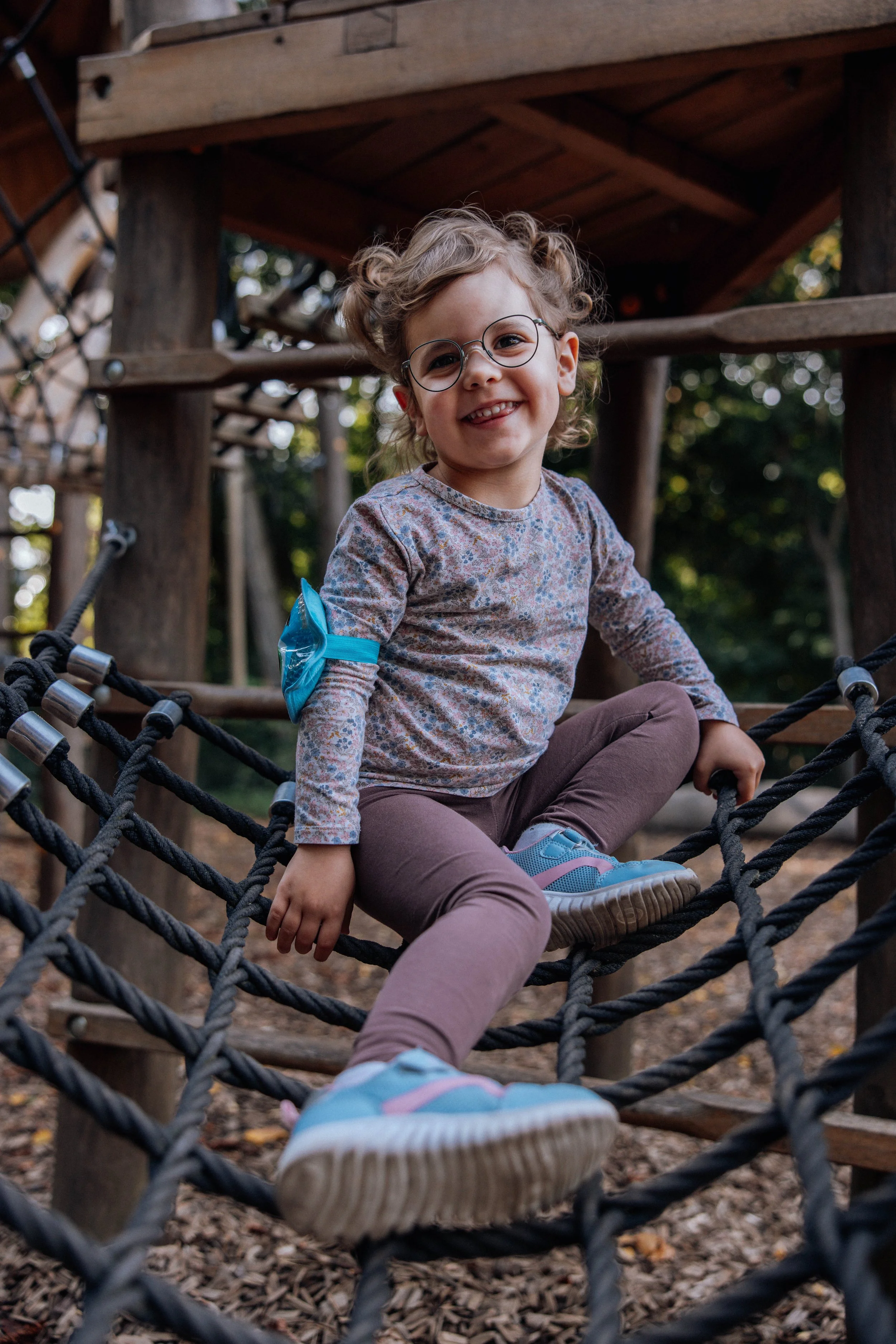 Ein lachendes Mädchen mit Brille, das auf einer Seilbrücke in einem Spielplatz sitzt.