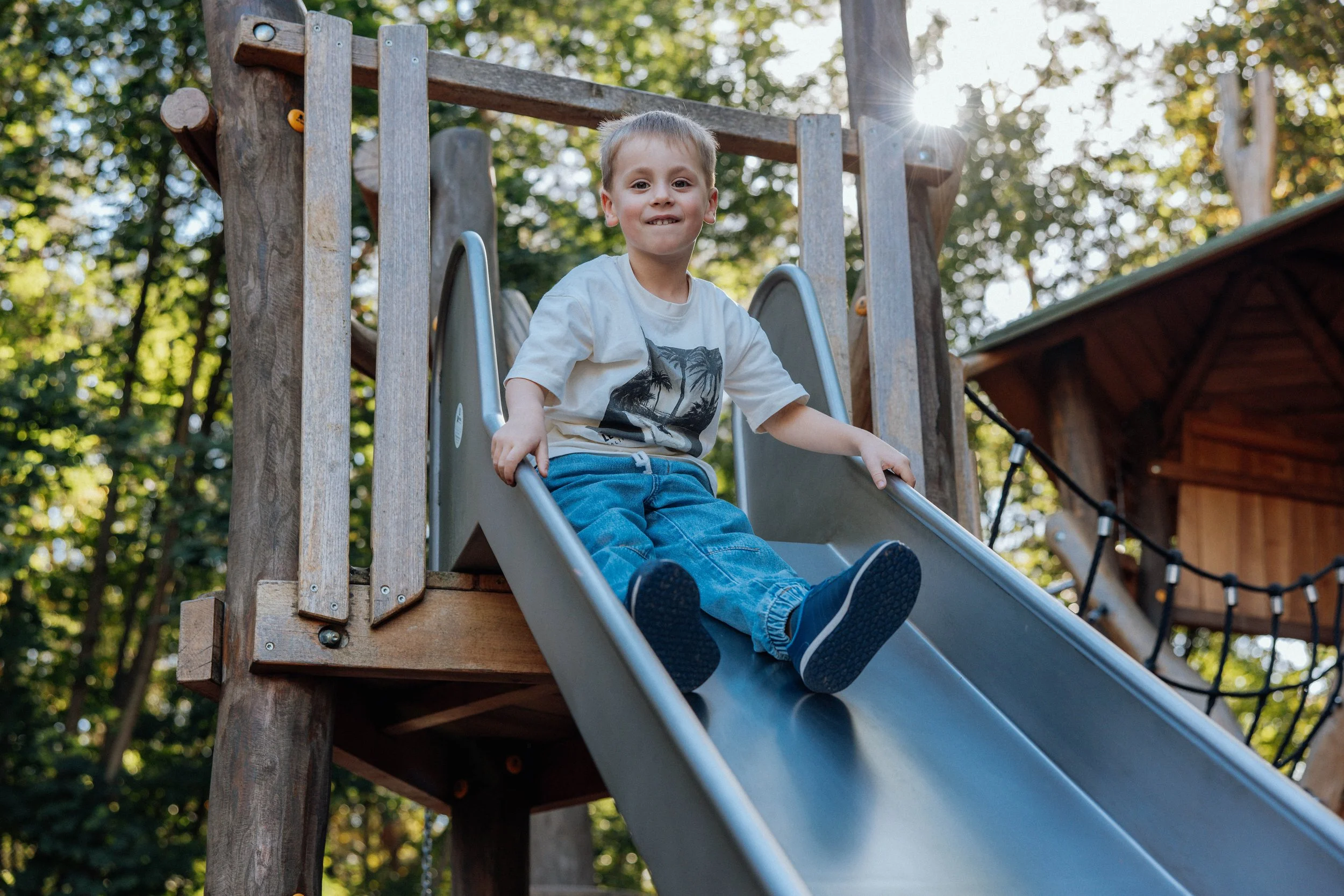Ein Junge sitzt auf einer Rutsche in einem Kinderspielplatz im Freien, umgeben von Bäumen mit Sonnenlicht im Hintergrund.
