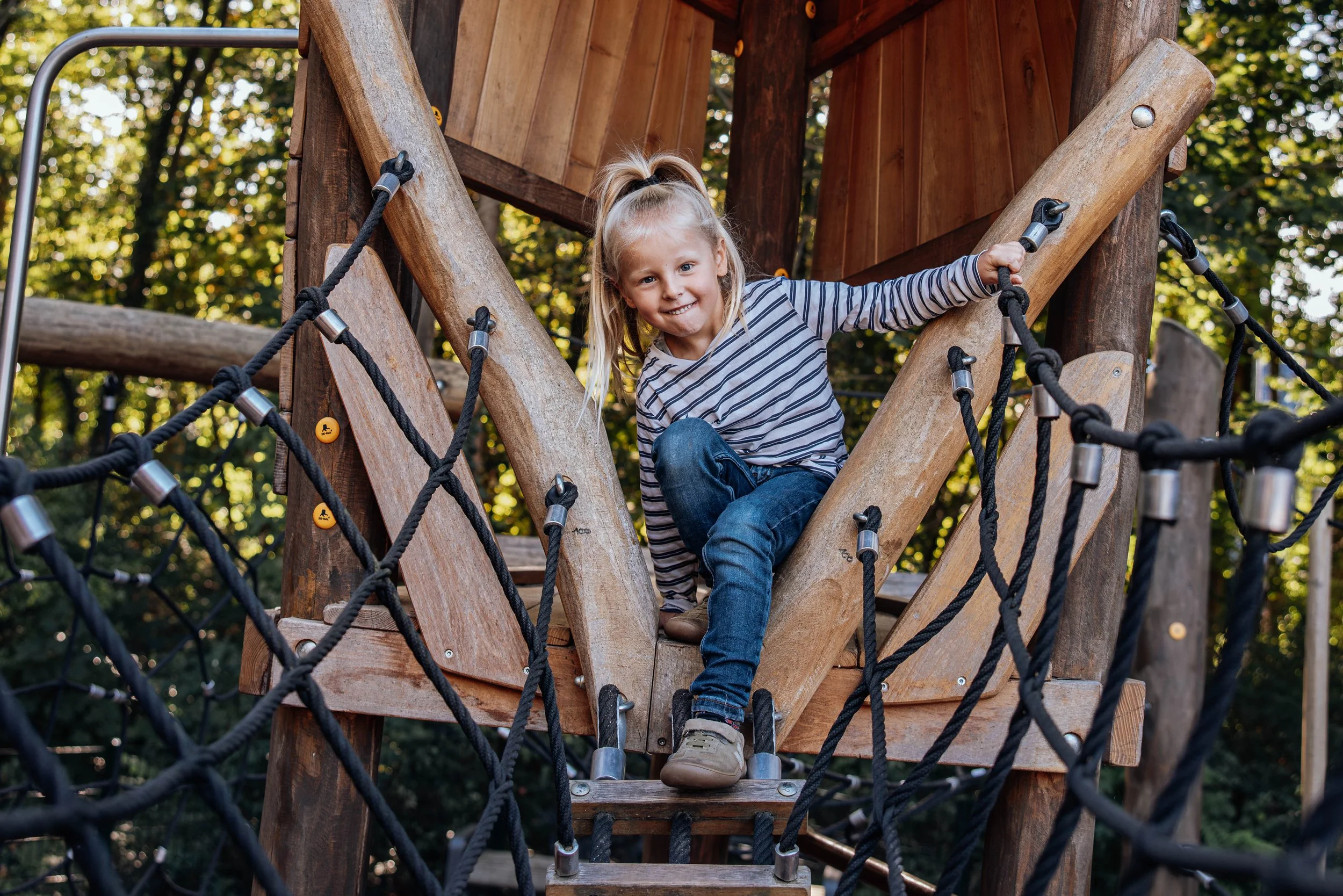 Ein Mädchen spielt auf einem Kletterspielplatz im Freien, umgeben von Bäumen, und lächelt in die Kamera.