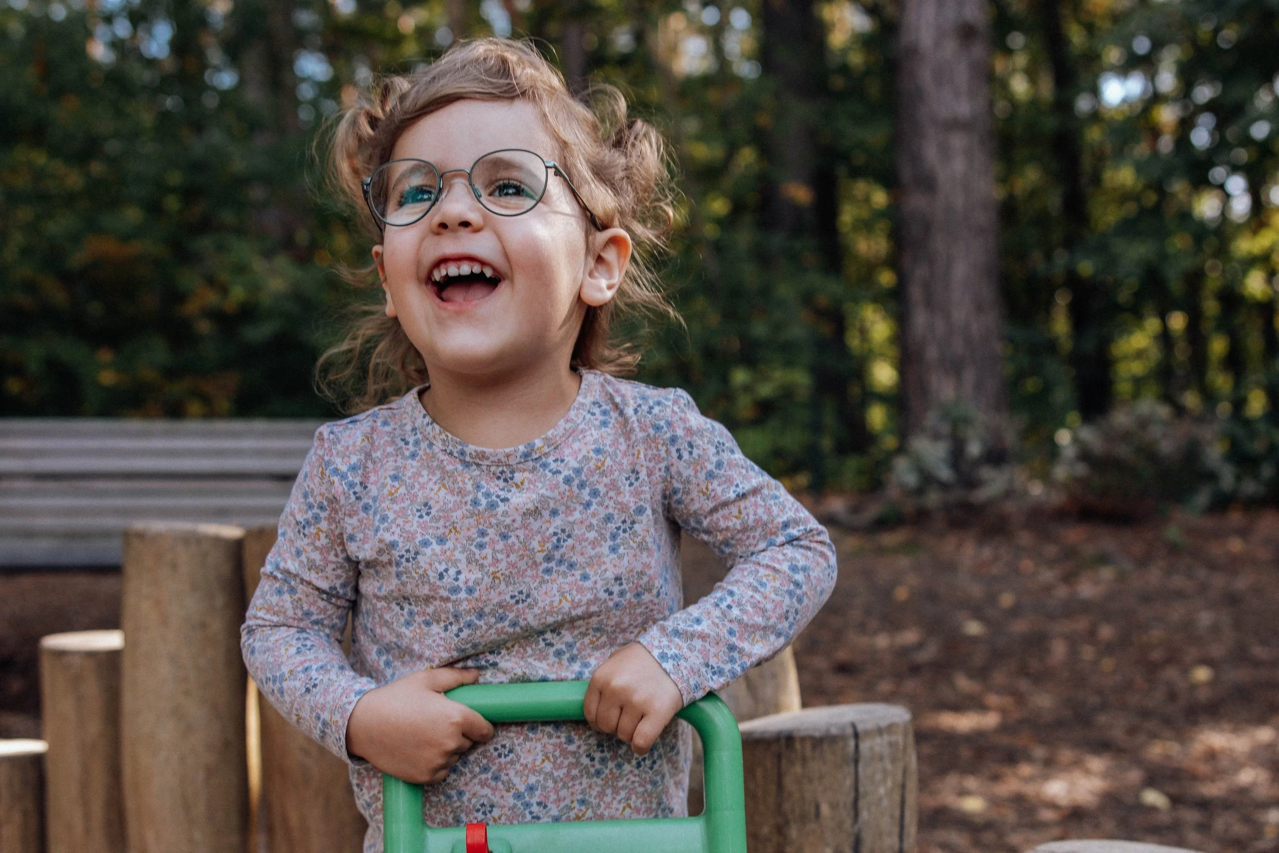 Lachendes kleines Mädchen mit Brille im Freien, umgeben von Bäumen, mit einem Spielzeug in der Kita in Berlin