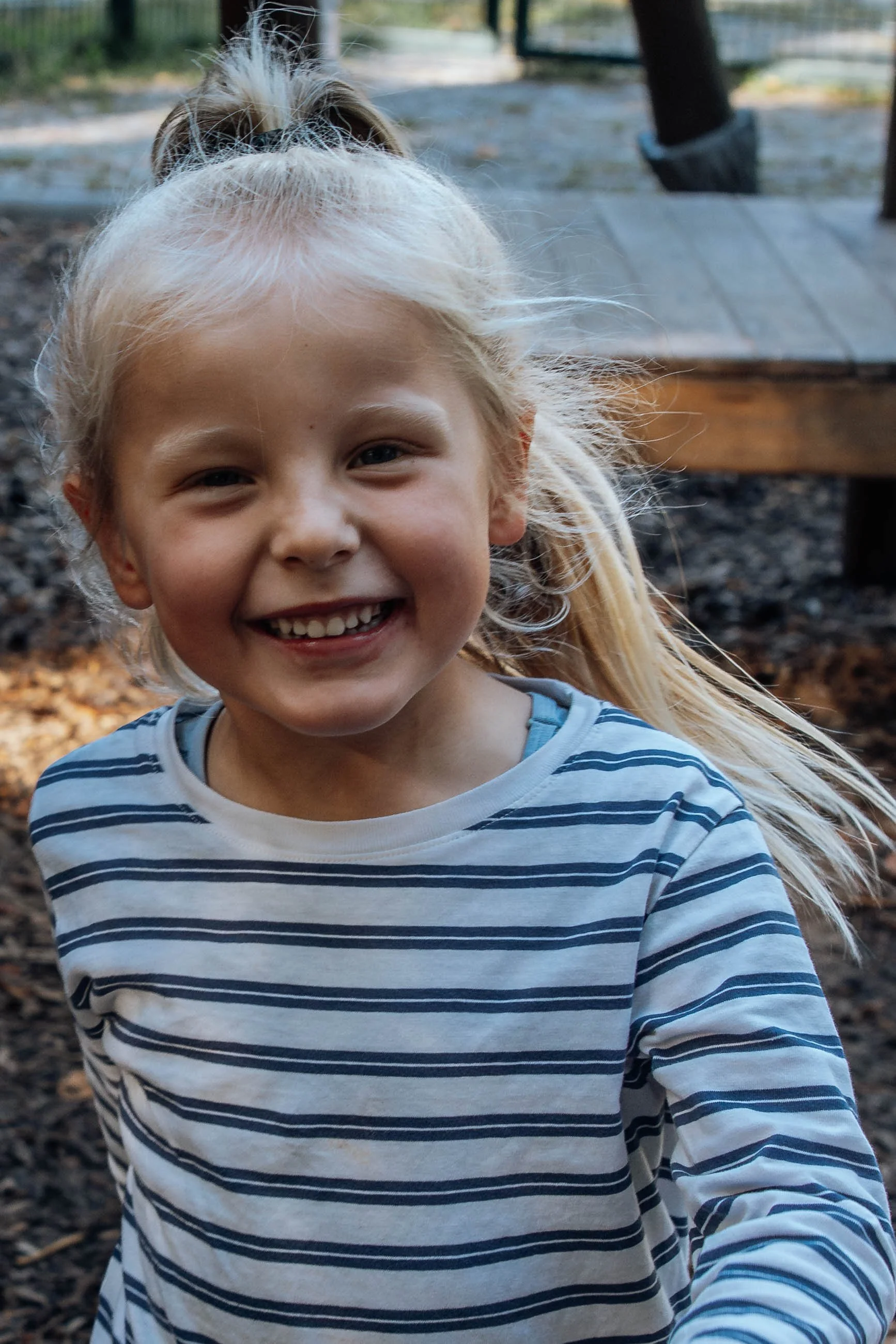Ein lachendes Mädchen mit blondem Haar, das einen blauen und weißen gestreiften Pullover trägt, draußen im Park bei Sonnenuntergang.
