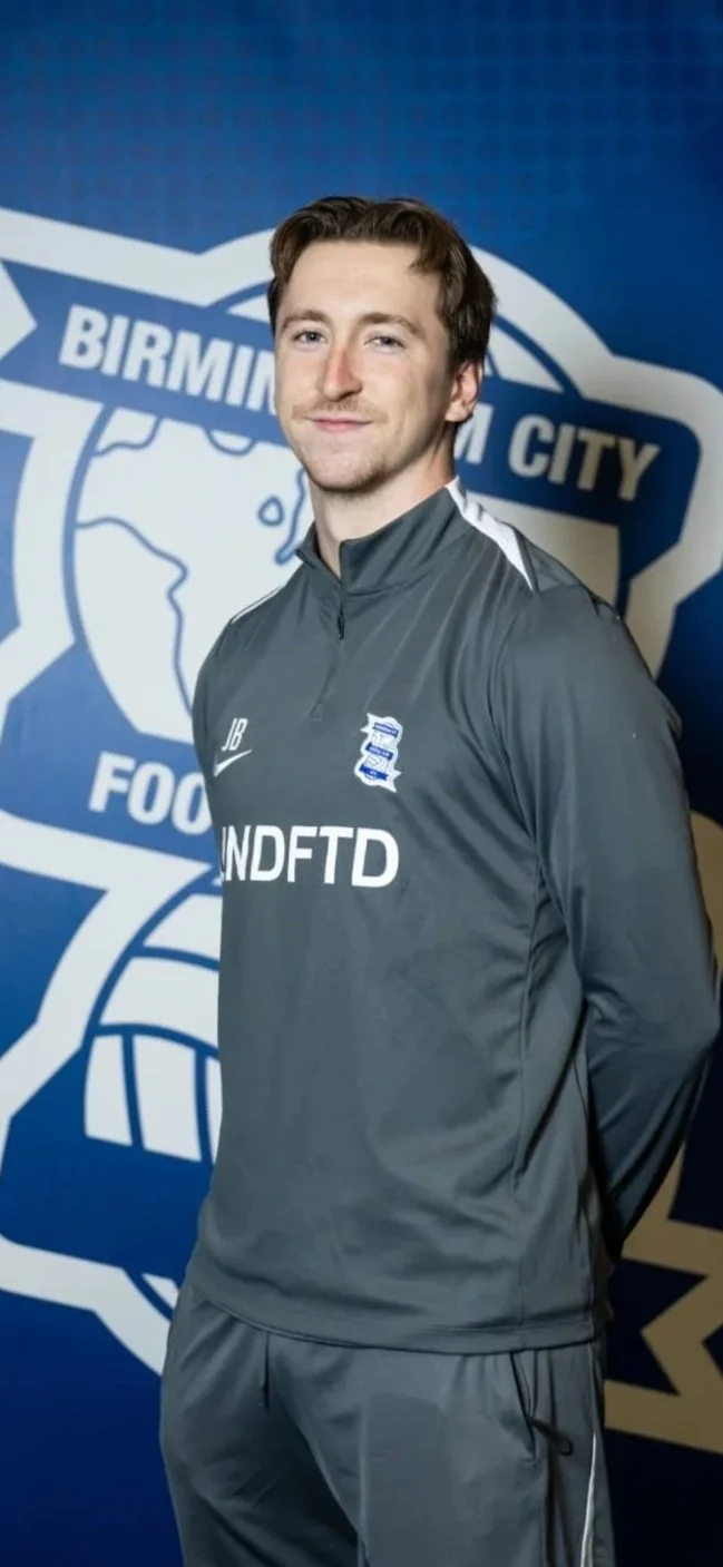 A young man with brown hair in sportswear standing in front of a Birmingham City background logo.