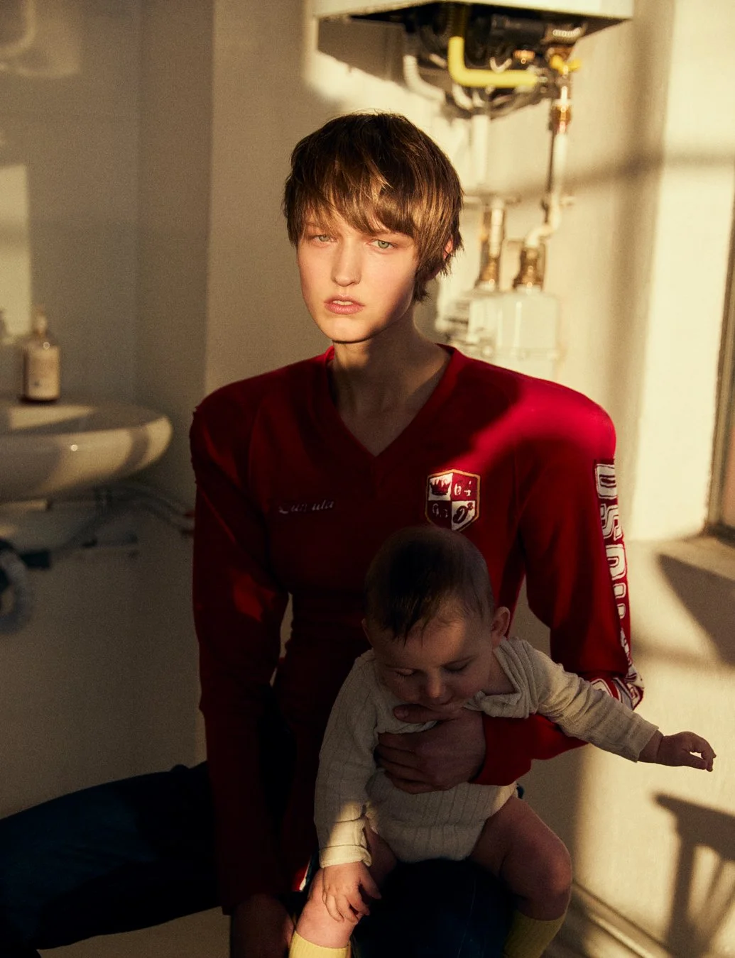 A young woman with short brown hair wearing a red sports jersey holding a toddler in a cream-colored hoodie, in a room with sunlight coming through a window.