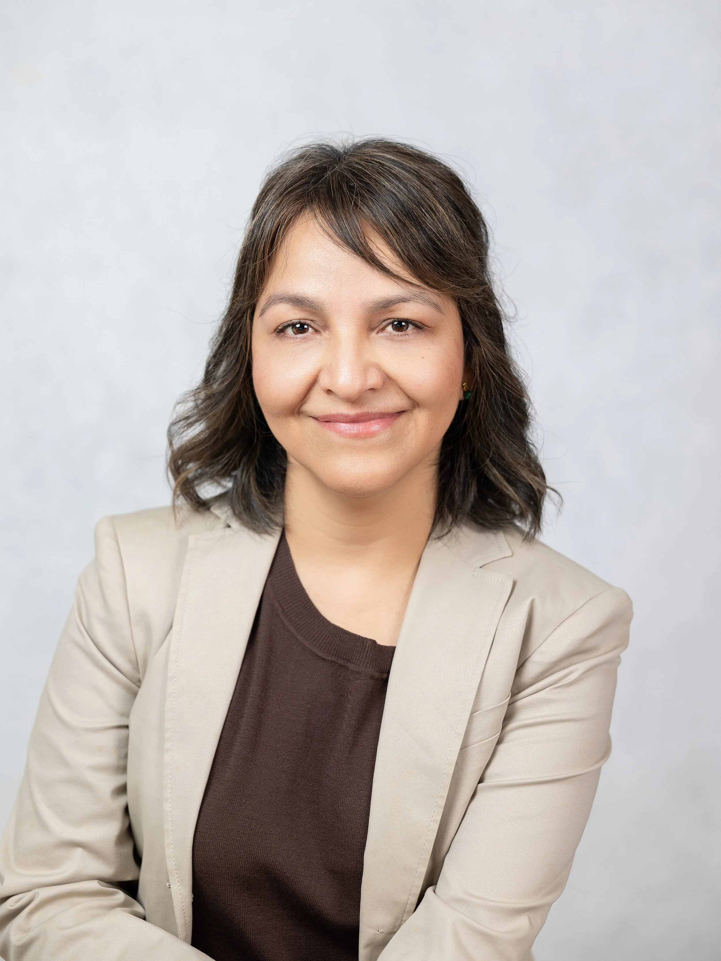 Mujer con cabello castaño y ondulado, vestida con blazer beige y blusa marrón, sonriendo frente a fondo gris claro.