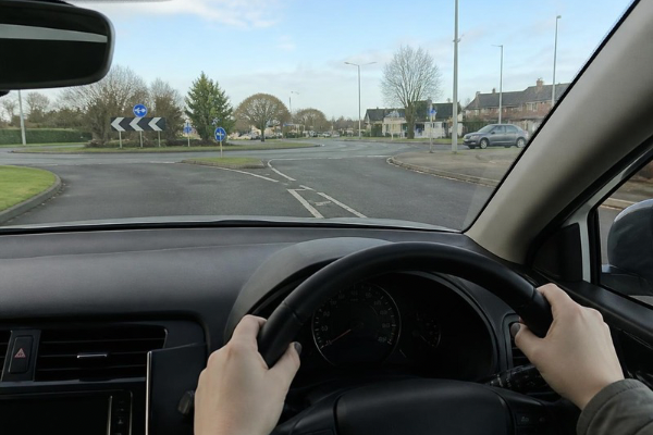 Inside a car, a person is holding the steering wheel at a roundabout with multiple signs indicating directions, trees, and houses in the background.
