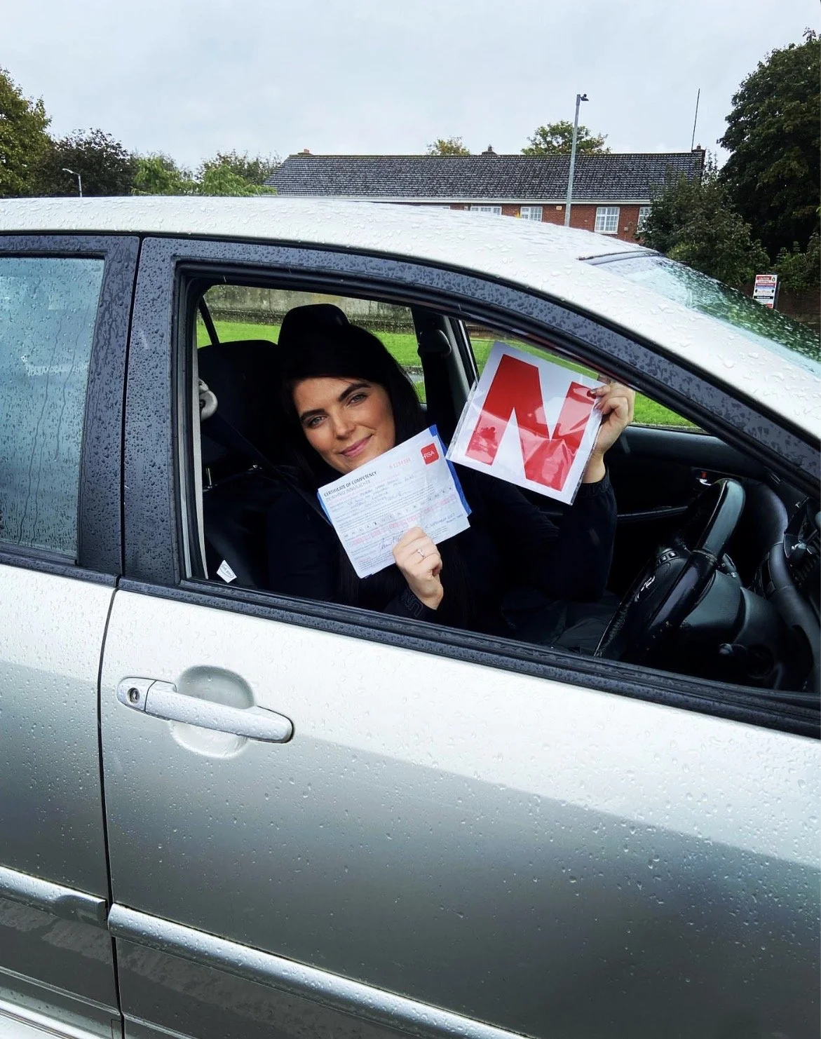 Woman sitting in a silver car holding a driving test pass letter and a driving exam fail sign, with raindrops on the car window.