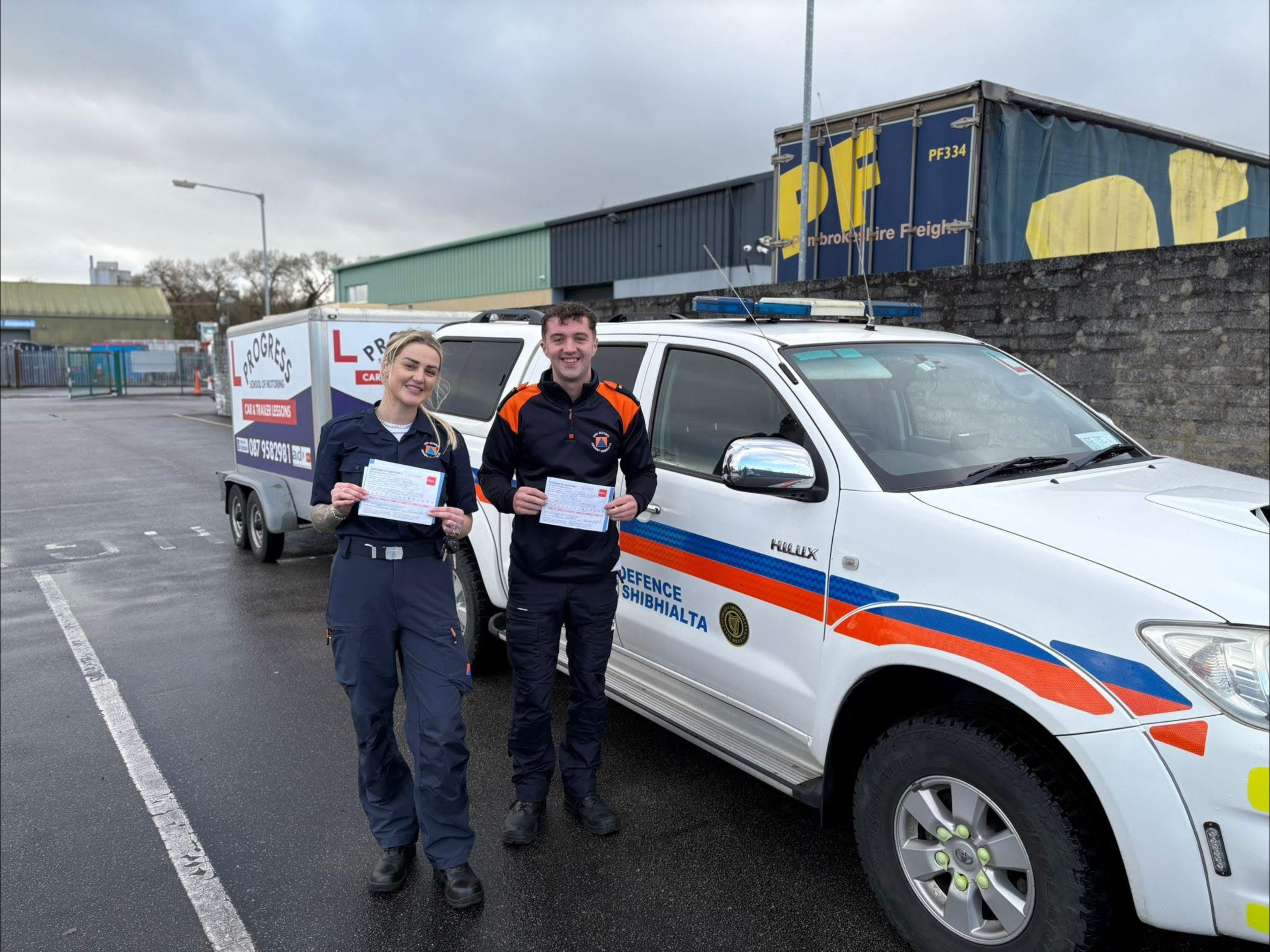 Two paramedics standing next to a white ambulance, holding documents, with a storage and cargo area nearby in an outdoor parking lot.