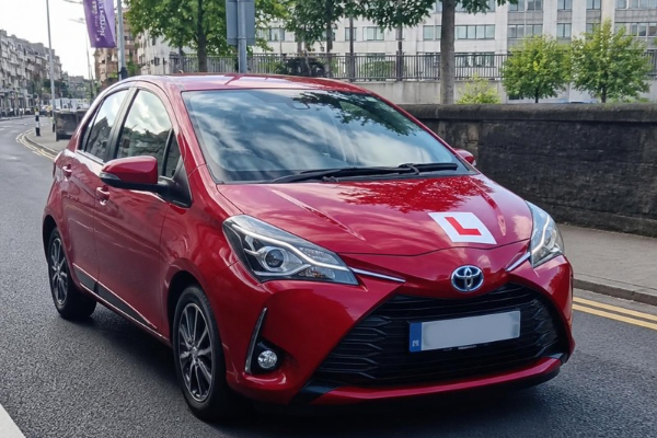 Red Toyota Corolla driving on city street with learner driver sign on hood.