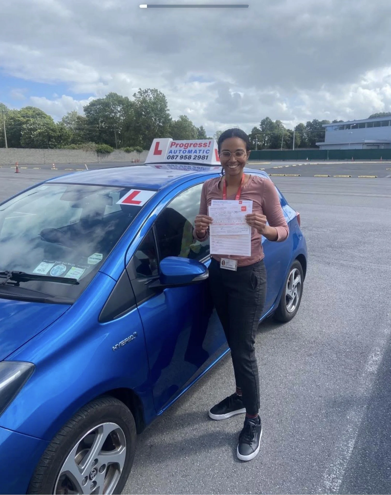 A woman standing next to a blue hybrid car holding a paper, smiling, in a parking lot with a driving school sign on top of the car that reads 'Progress! Automatic' and includes a phone number, with trees and a building in the background.