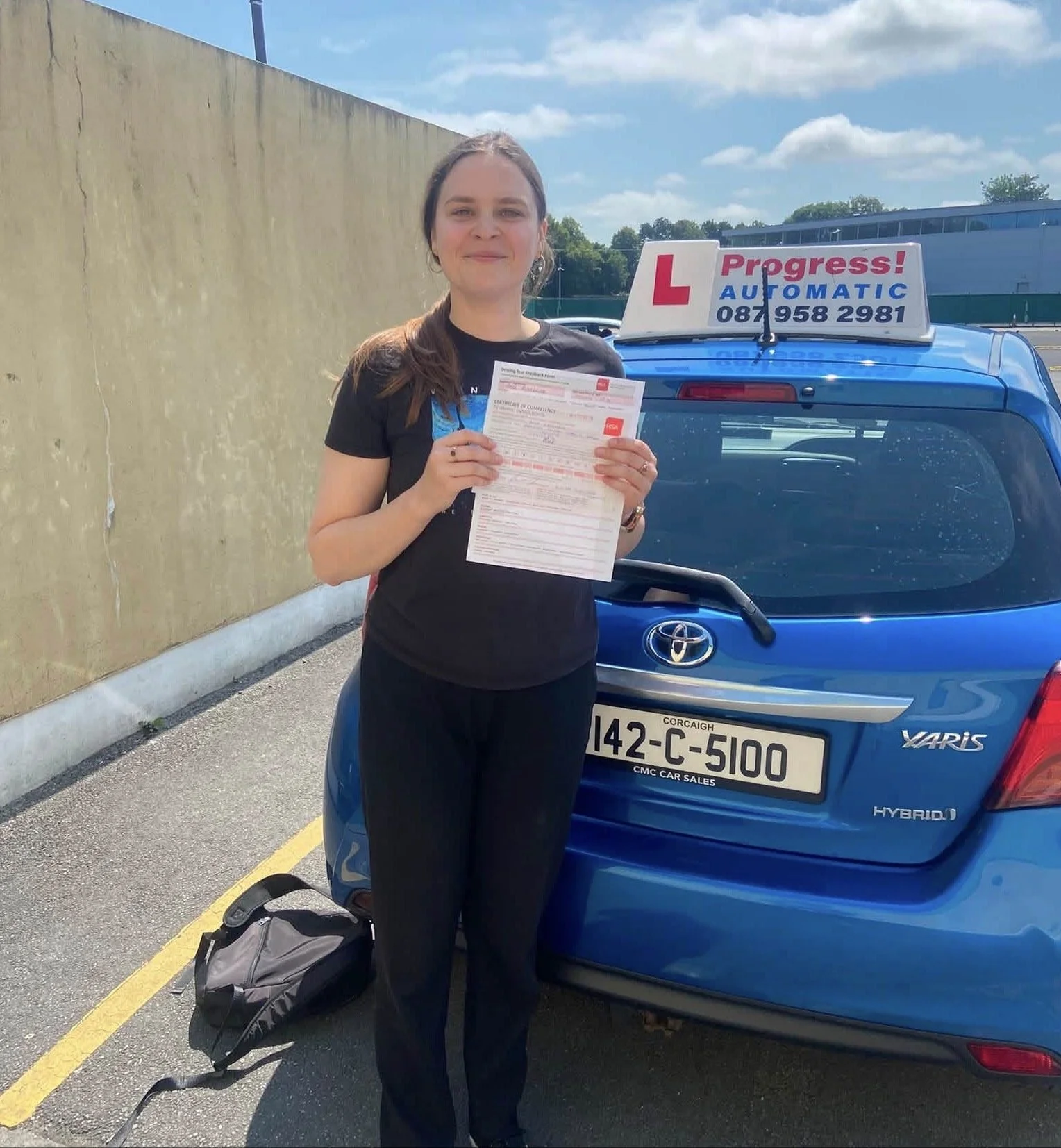 Woman holding driving test paperwork standing next to a blue hybrid Toyota Yaris with a driving school sign on top. She has a backpack on the ground nearby, a yellow line on the pavement, and a beige wall on the left. Cloudy blue sky overhead.