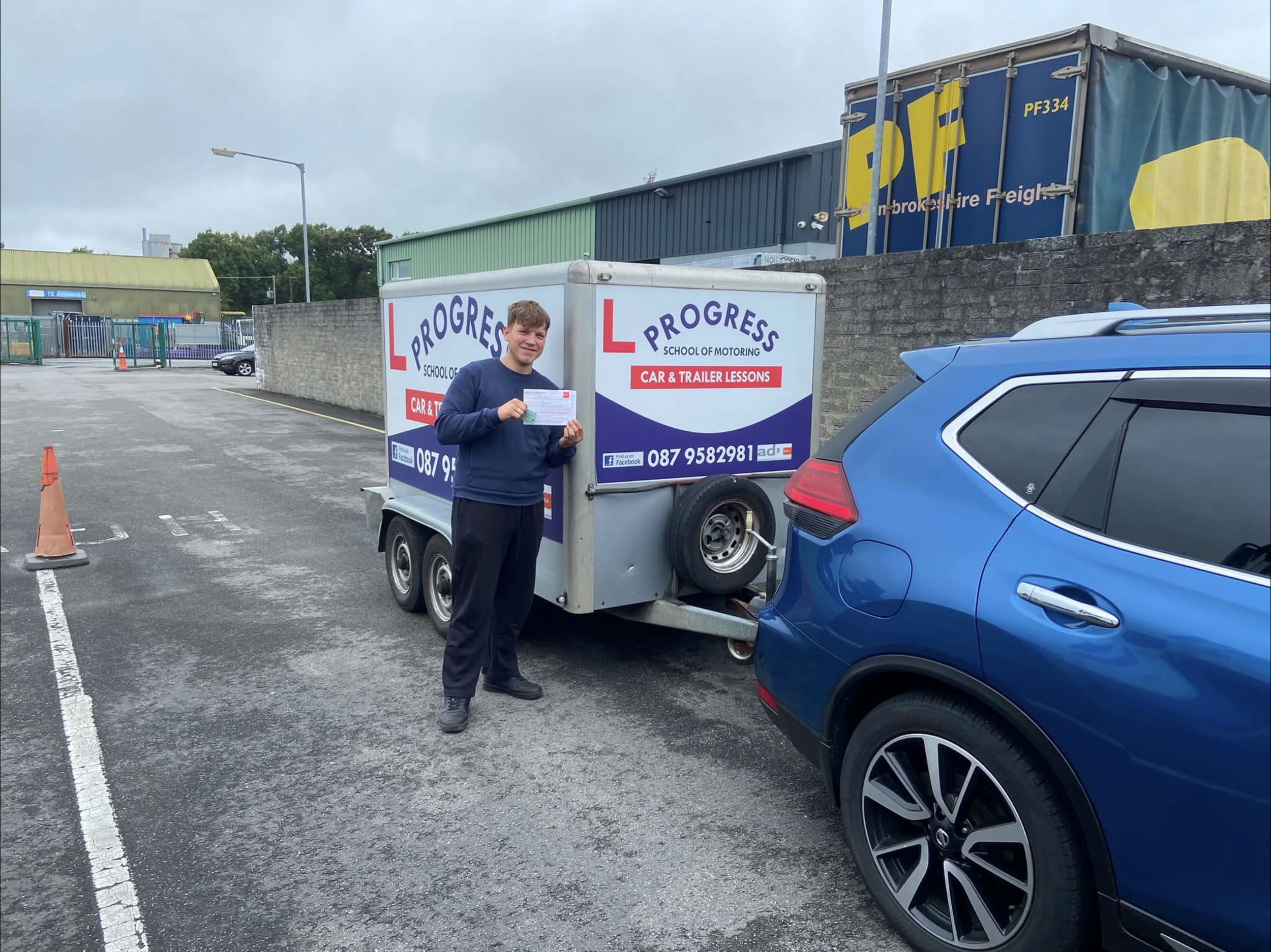 A man standing in a parking lot holding a paper, next to a trailer for a driving school called Progress School of Motoring with car and trailer lessons advertisement, with cars and industrial buildings in the background.