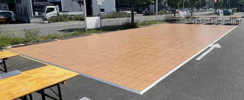 Eine große, quadratische, braune, tileartige Fläche, vermutlich eine markierte Zone für Veranstaltungen, mit Bänken und Tischen im Hintergrund auf einer Straße in einer städtischen Umgebung.