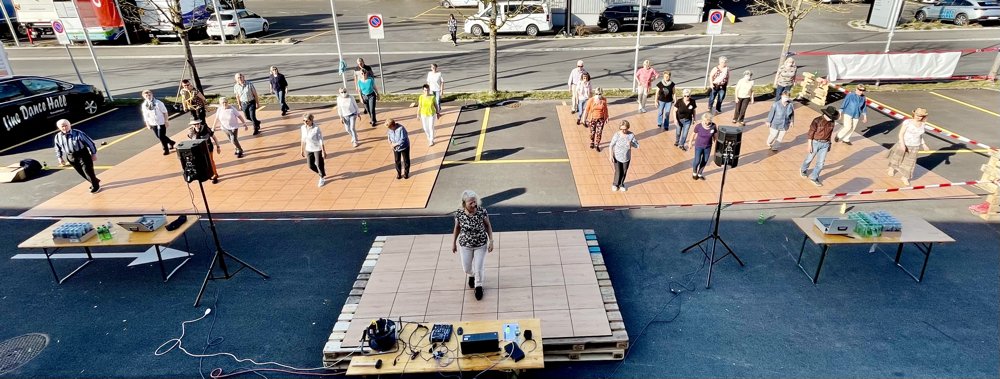 Menschen tanzen auf zwei abgetrennten Tanzflächen im Außenbereich, mit einer Person in der Mitte auf einer kleinen Bühne. Tische mit Getränken und Technik stehen neben den Tanzflächen, im Hintergrund sind parkende Autos und Bäume sichtbar.