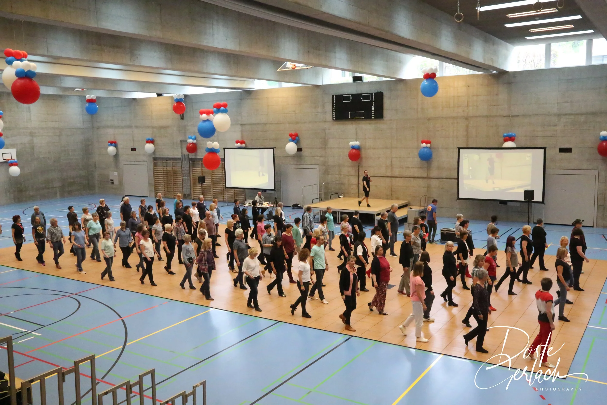 Menschen versammeln sich in einem Turnhalle für eine Veranstaltung, mit Ballons an der Decke und zwei große Bildschirme an den Wänden.