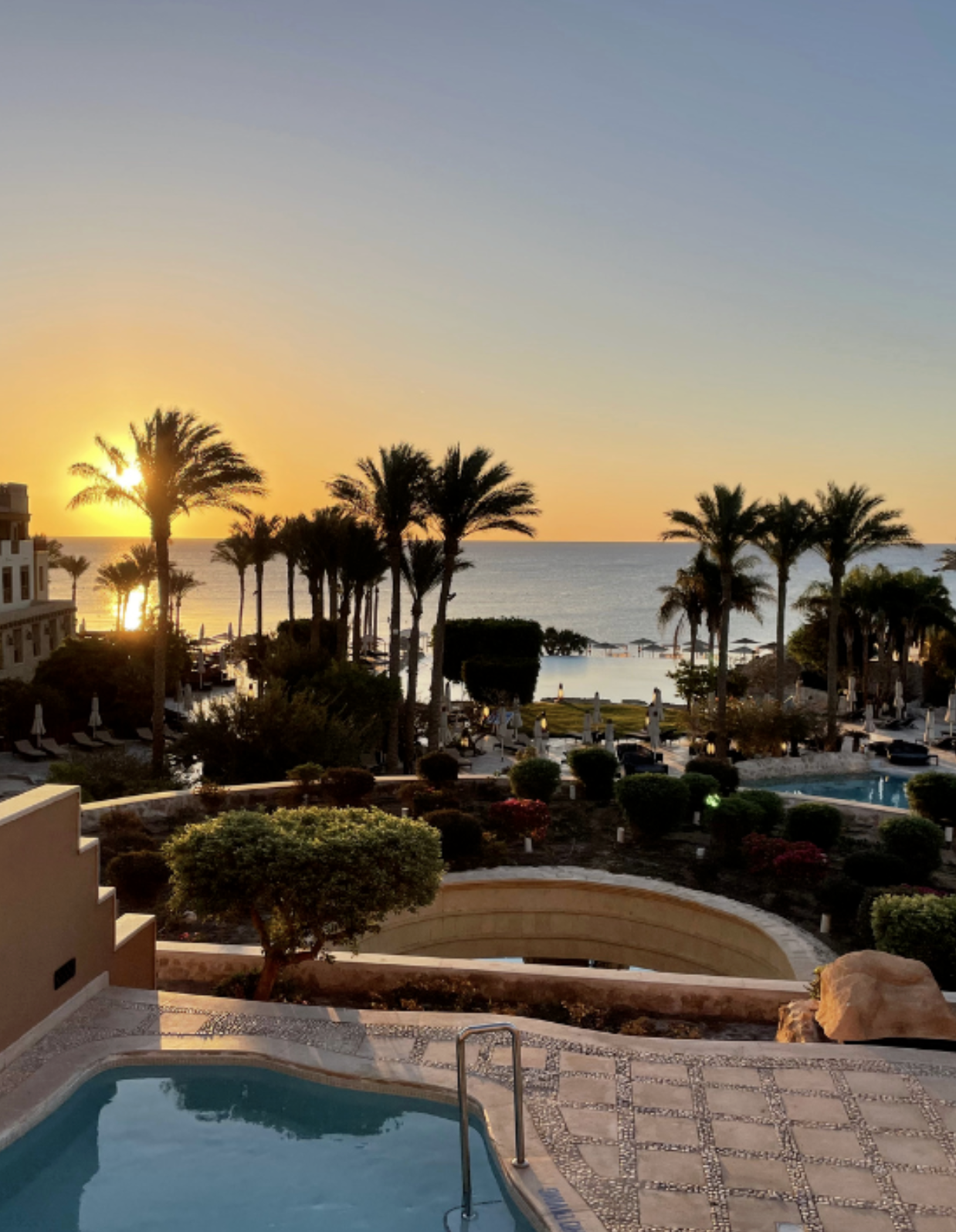 Sonnenuntergang über einem Pool mit Palmen, Meer im Hintergrund, Liegestühlen und Sonnenschirme am Strand.