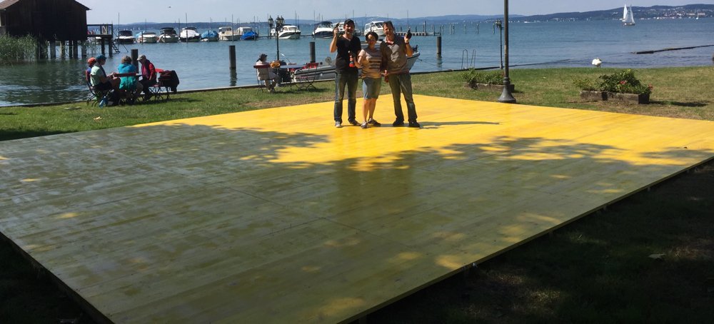 Drei Menschen stehen auf einer großen gelben Plattform im Park, im Hintergrund ist ein Wasserarm mit Segelbooten und Menschen, die am Ufer sitzen.