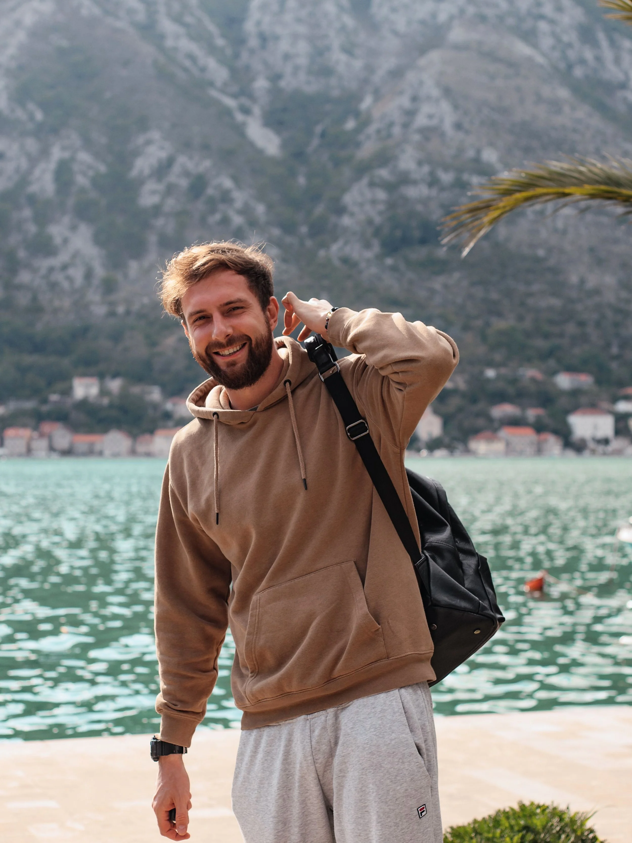 A smiling man with a beard and casual clothing, standing near water with mountains in the background.