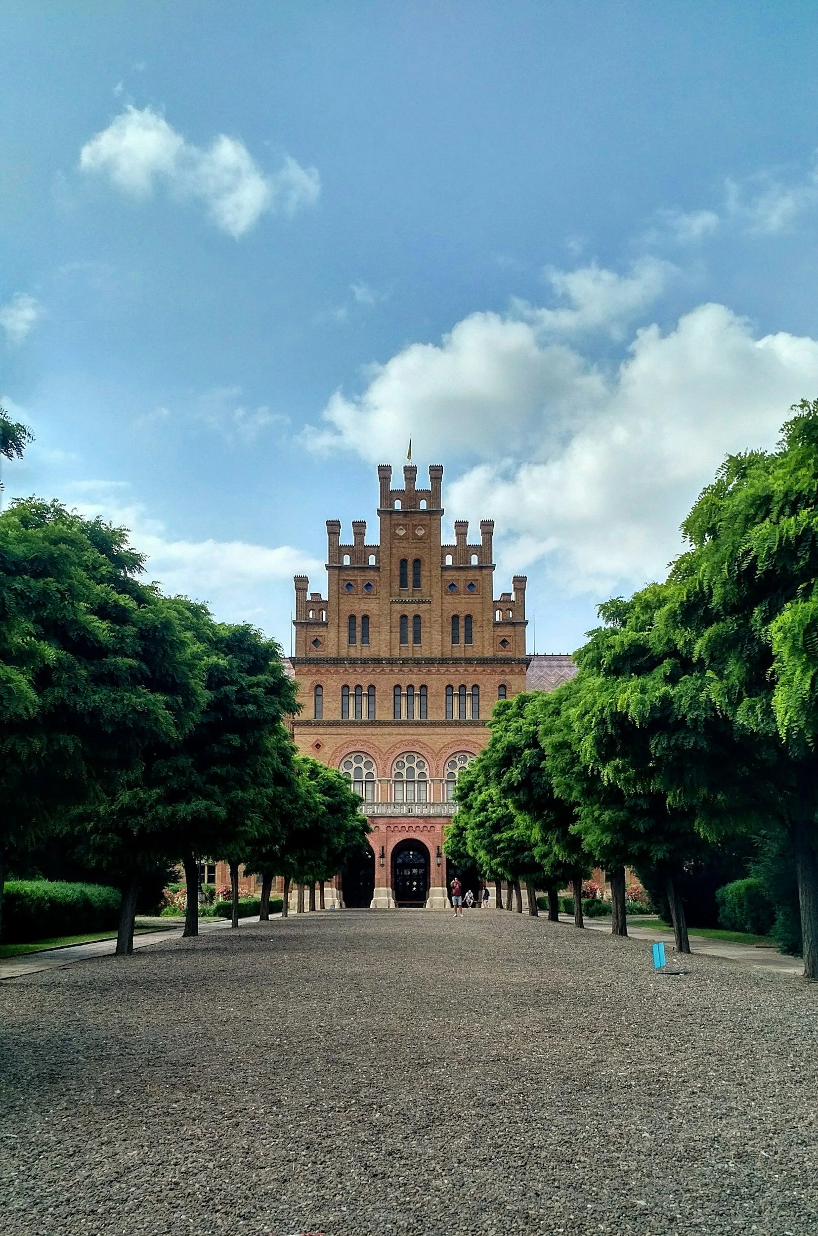 A historic brick castle-like building with a flag on top, framed by lush green trees on a gravel pathway, under a blue sky with scattered clouds.