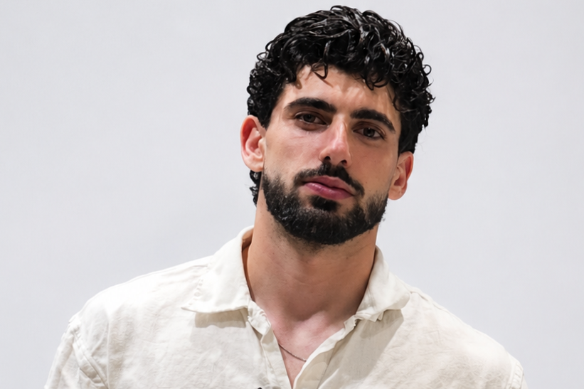 A young man with curly dark hair and a beard wearing a light-colored button-up shirt against a plain background.