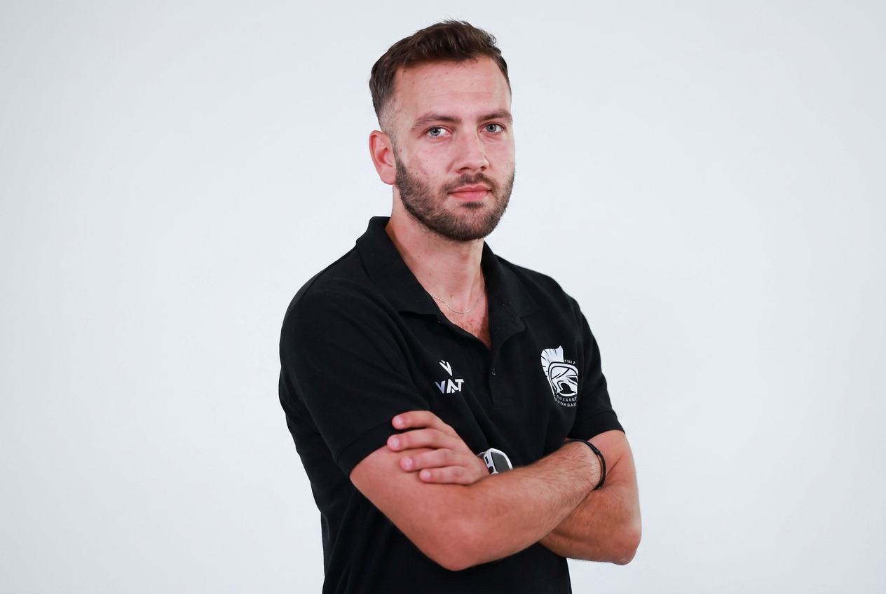 Young man with crossed arms, wearing a black polo shirt with logos, standing against a plain white background.