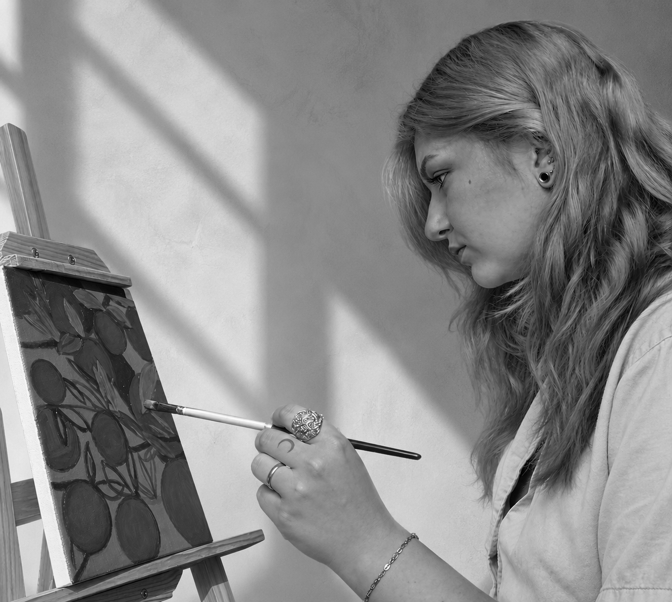 A woman with wavy hair painting on a small canvas on an easel, using a brush, with sunlight casting shadows on the wall behind her.