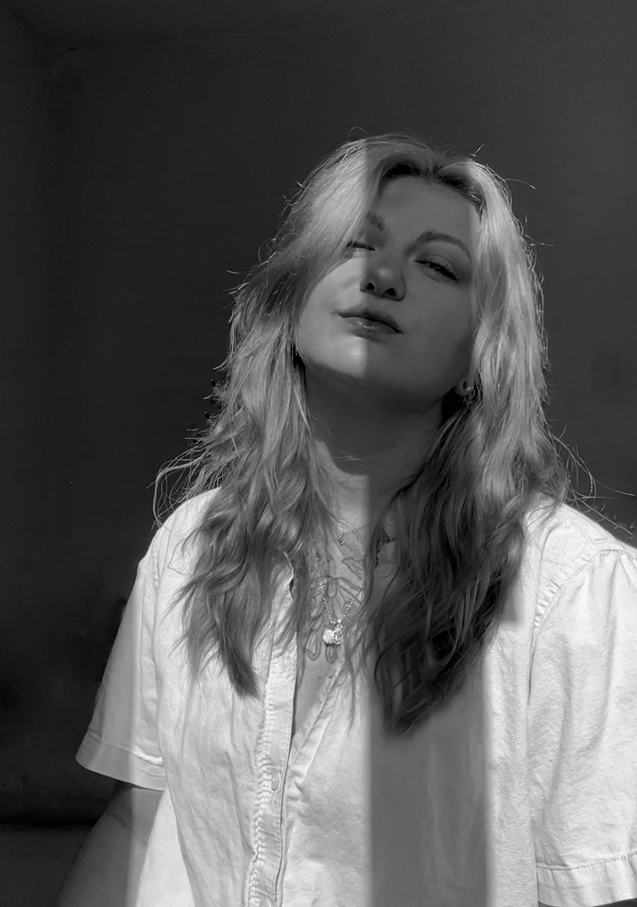 A black and white portrait of a young woman with wavy blonde hair, wearing a white shirt, looking at the camera with a confident expression, with light falling on her face.