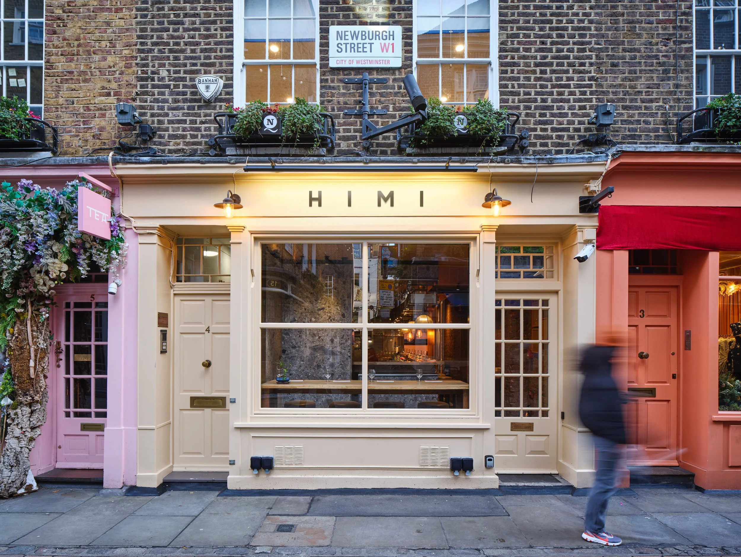 Storefront with cream-colored awning that has the word 'HIMI' displayed, large window, brick building above with potted plants, street sign for Newburgh Street W1, pink building to the left with floral decoration, pink door to the right, and a blurred person walking by.