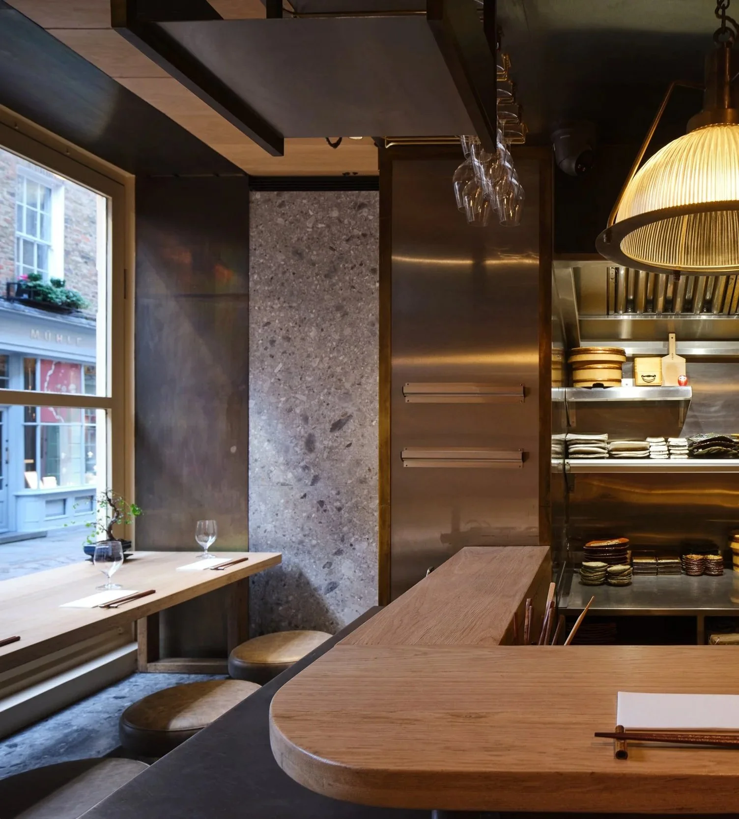Interior of a restaurant with wooden tables, glasses set on the tables, a window showing buildings outside, and shelves with dishes and glassware.